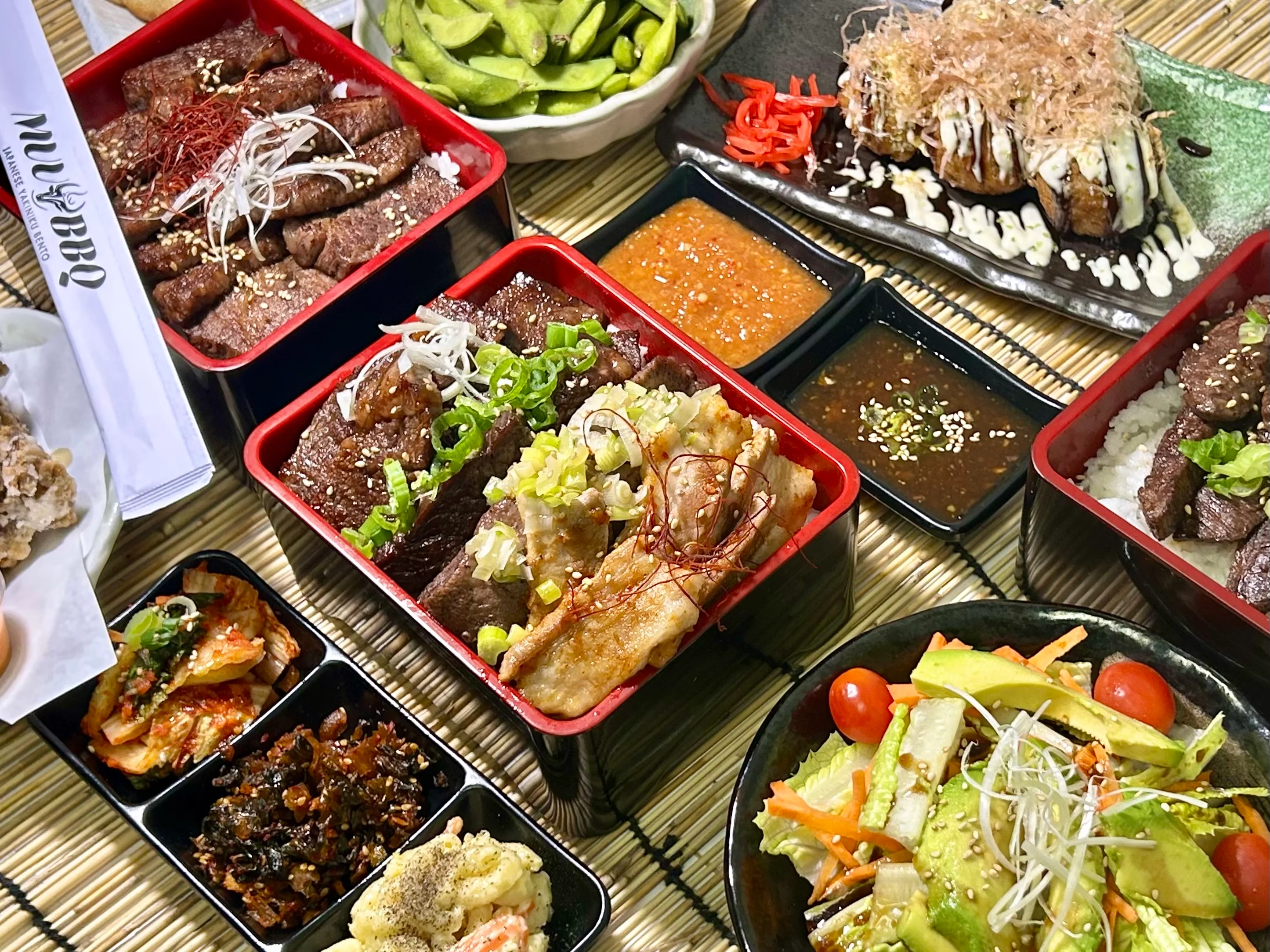 An assortment of Japanese dishes including bento boxes filled with grilled meats, rice, and various sides such as salad, pickles, and sauces. The meal is presented on a textured bamboo mat.