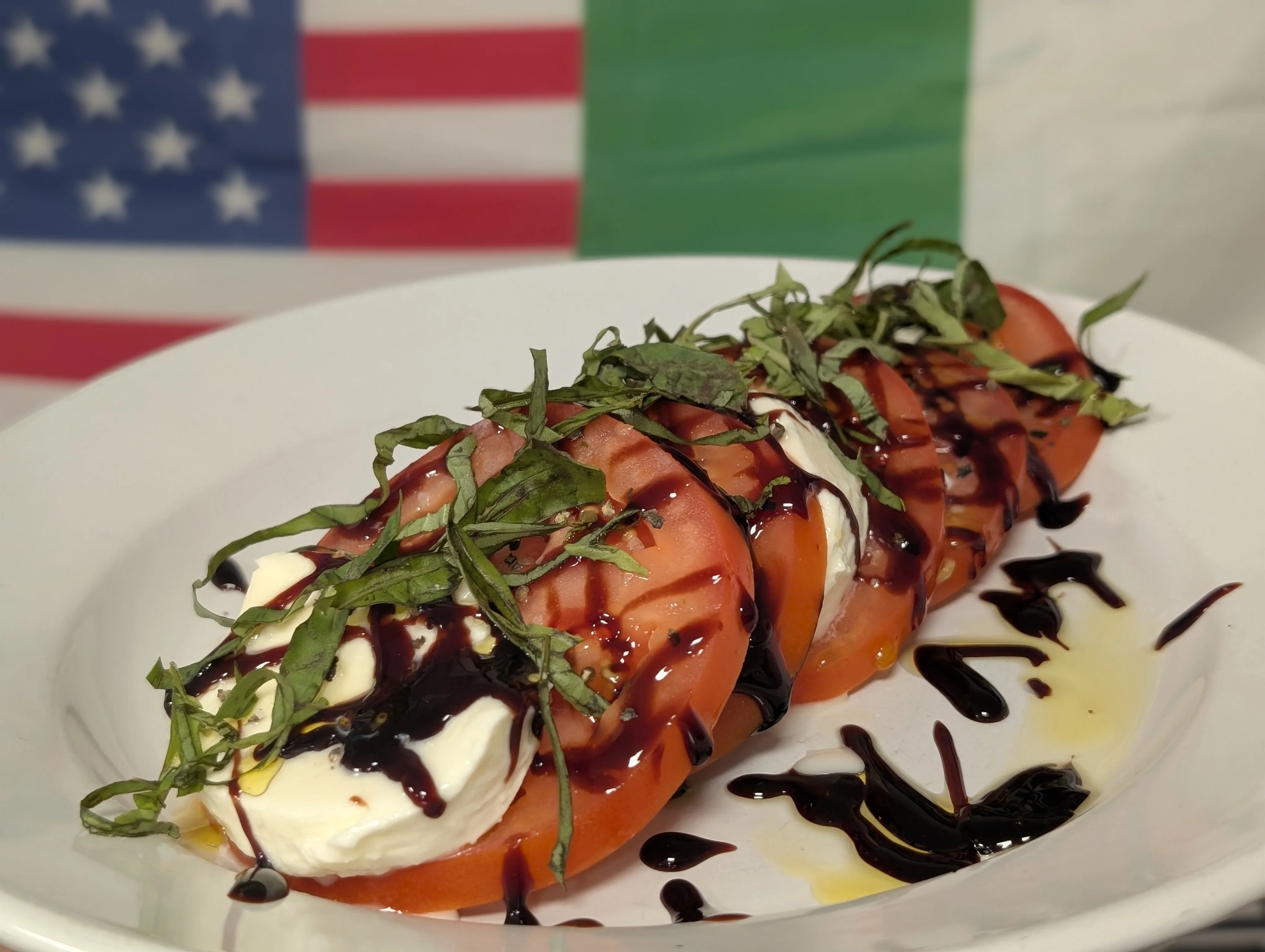A plate of fresh caprese salad featuring slices of tomato, mozzarella cheese, and fresh basil drizzled with balsamic reduction. The dish is set against a backdrop of flags.
