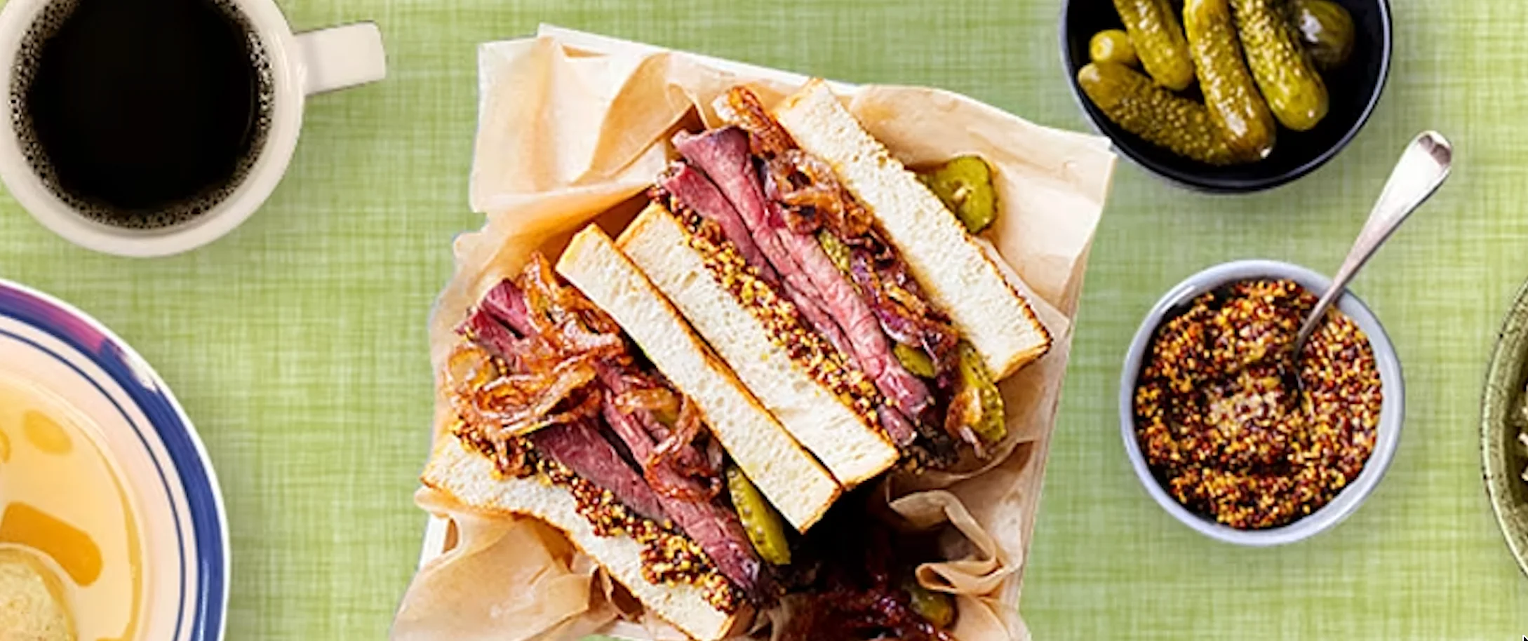 A sandwich filled with sliced meat and mustard, served with pickles and a cup of coffee, displayed over a green background.