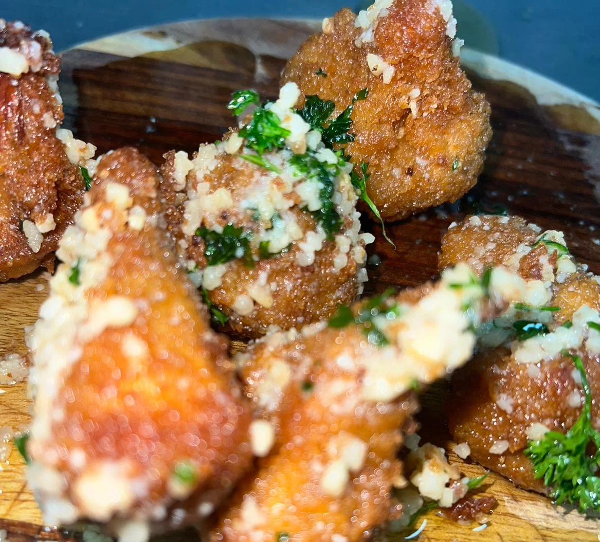 A close-up of golden fried balls, coated with herbs and topped with crushed nuts, served on a wooden plate.