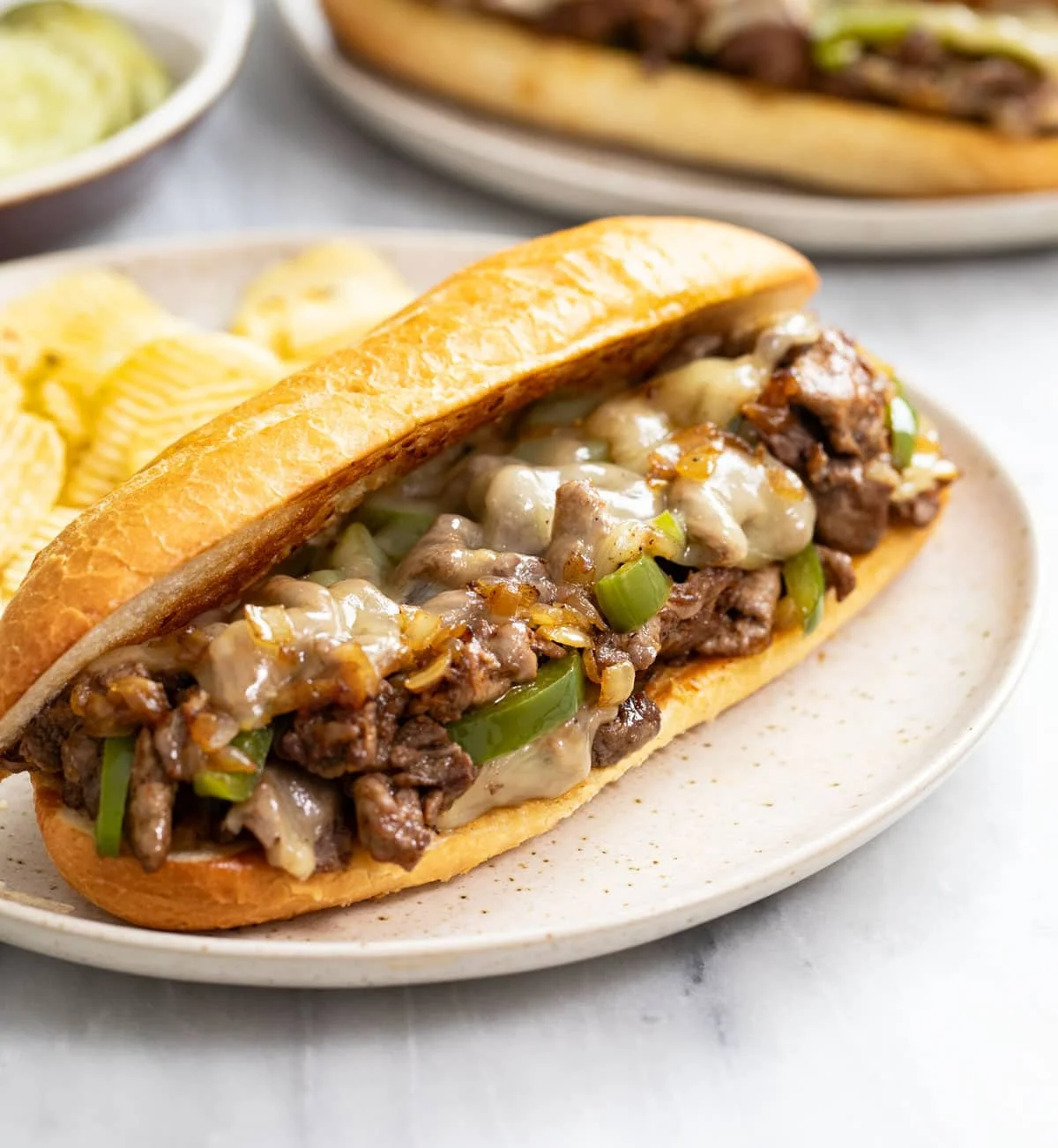 A soft hoagie filled with sautéed beef, melted cheese, and green bell peppers, served with a side of potato chips.