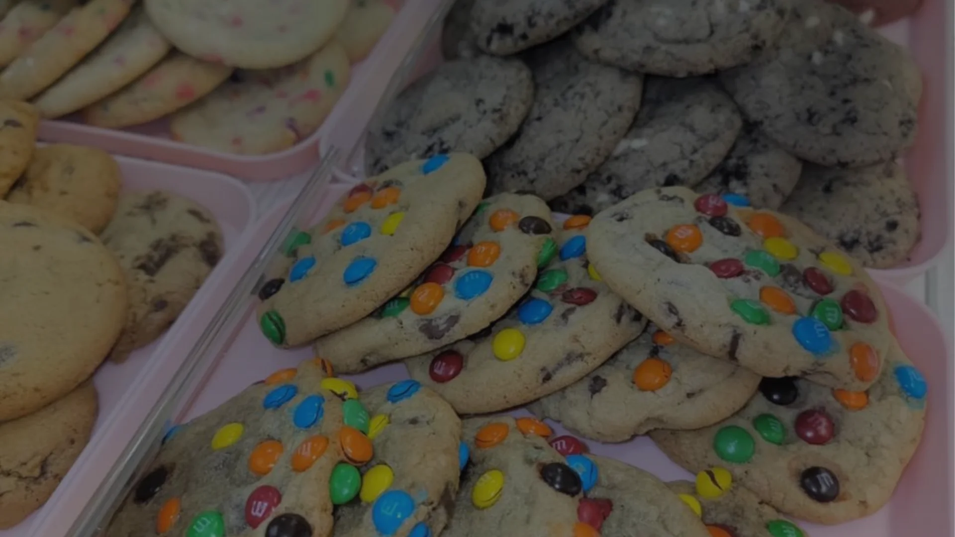 Trays of assorted cookies, including cookies topped with colorful M&M candies, chocolate chip cookies, and Oreo-style cookies.