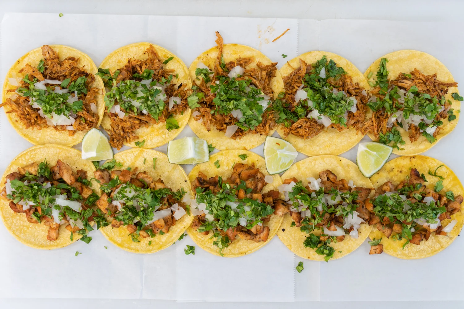 A close-up view of a lineup of tacos on parchment paper, filled with shredded meat, topped with cilantro and onions, and garnished with lime wedges.