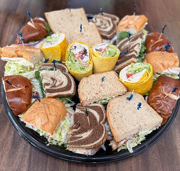 A platter of assorted sandwiches and wraps arranged on a black tray, featuring a variety of bread types including rye, whole wheat, pretzel rolls, and cheddar wraps. The sandwiches are filled with lettuce, meats, and other fresh ingredients, secured with blue toothpicks for easy serving.