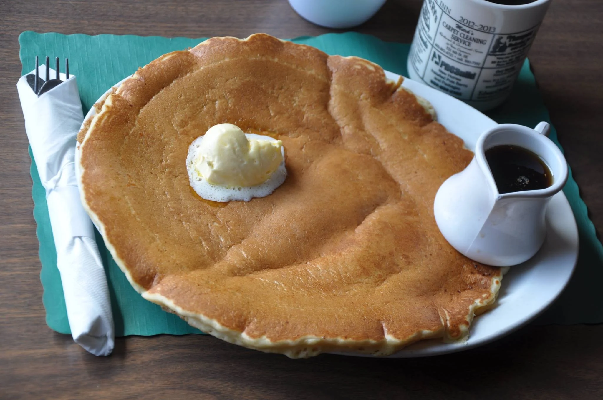 A large pancake topped with a scoop of butter, served with a small jug of syrup on a white plate.