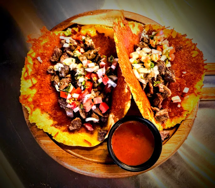A wooden platter holding two tacos filled with seasoned beef, fresh onions, and diced tomatoes, accompanied by a small bowl of sauce.