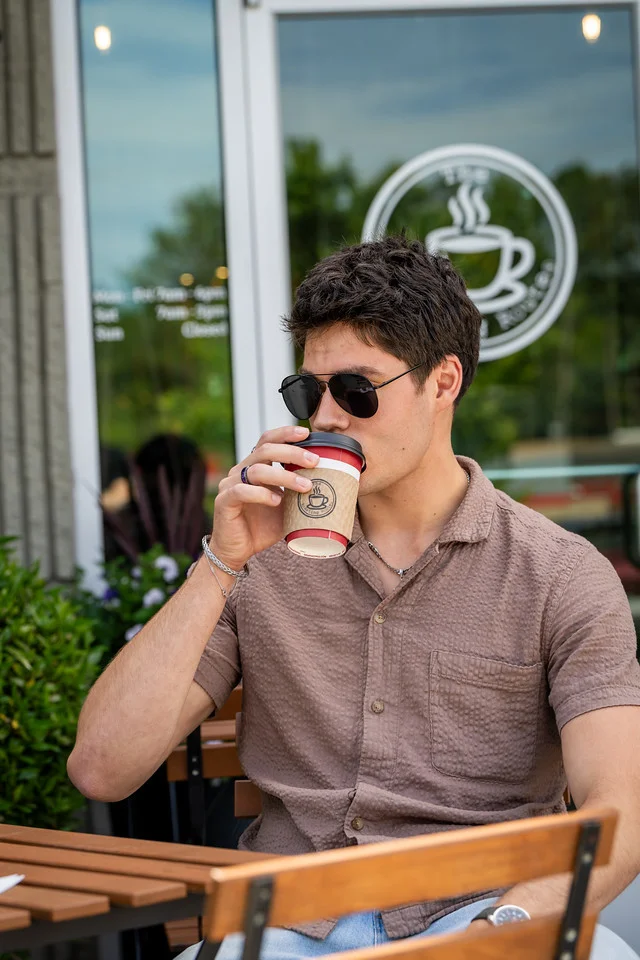 A young individual sitting outside a café, enjoying a beverage while wearing sunglasses. The setting includes a backdrop of greenery.