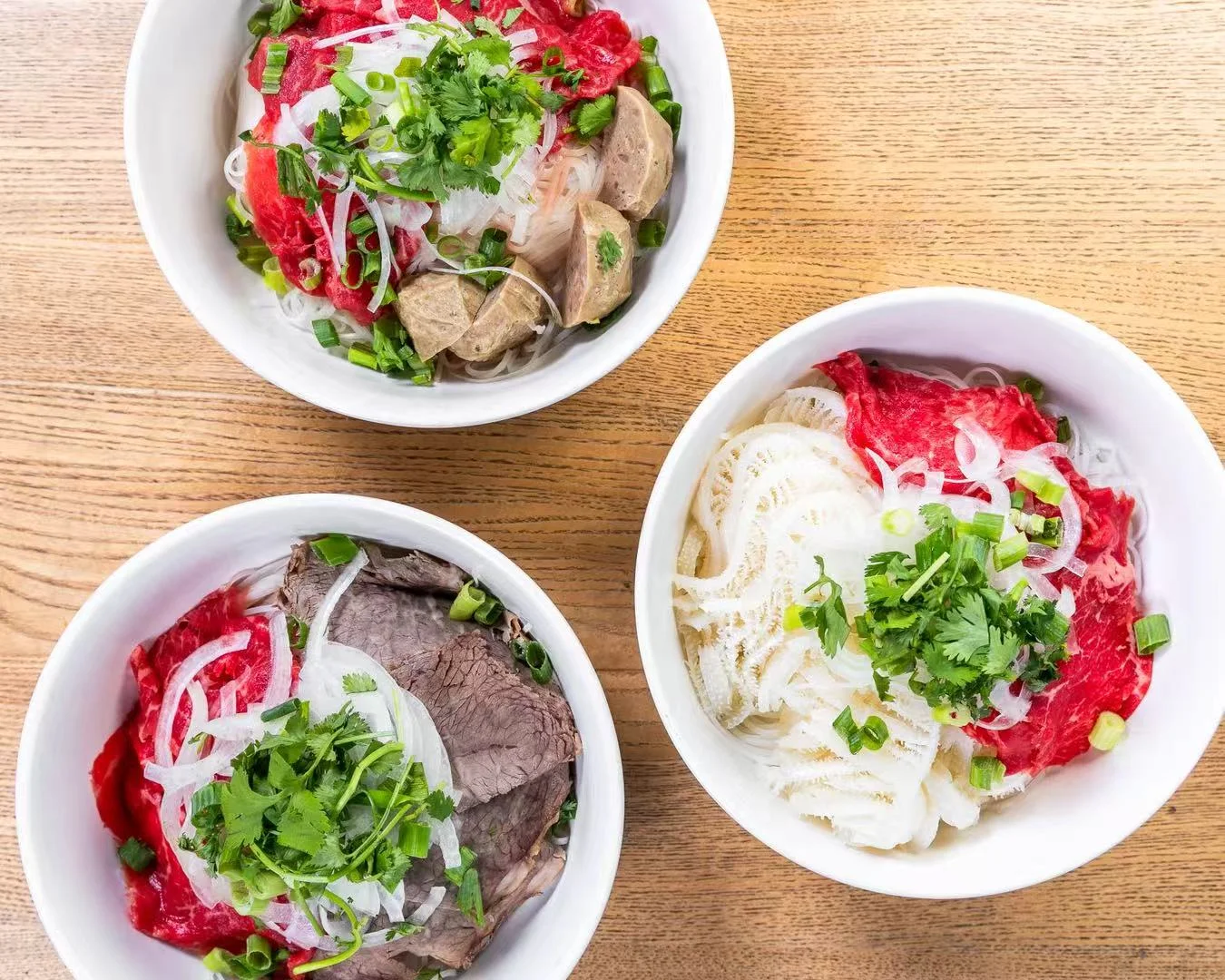 Three bowls of pho with various meats and garnishes on a wooden table.