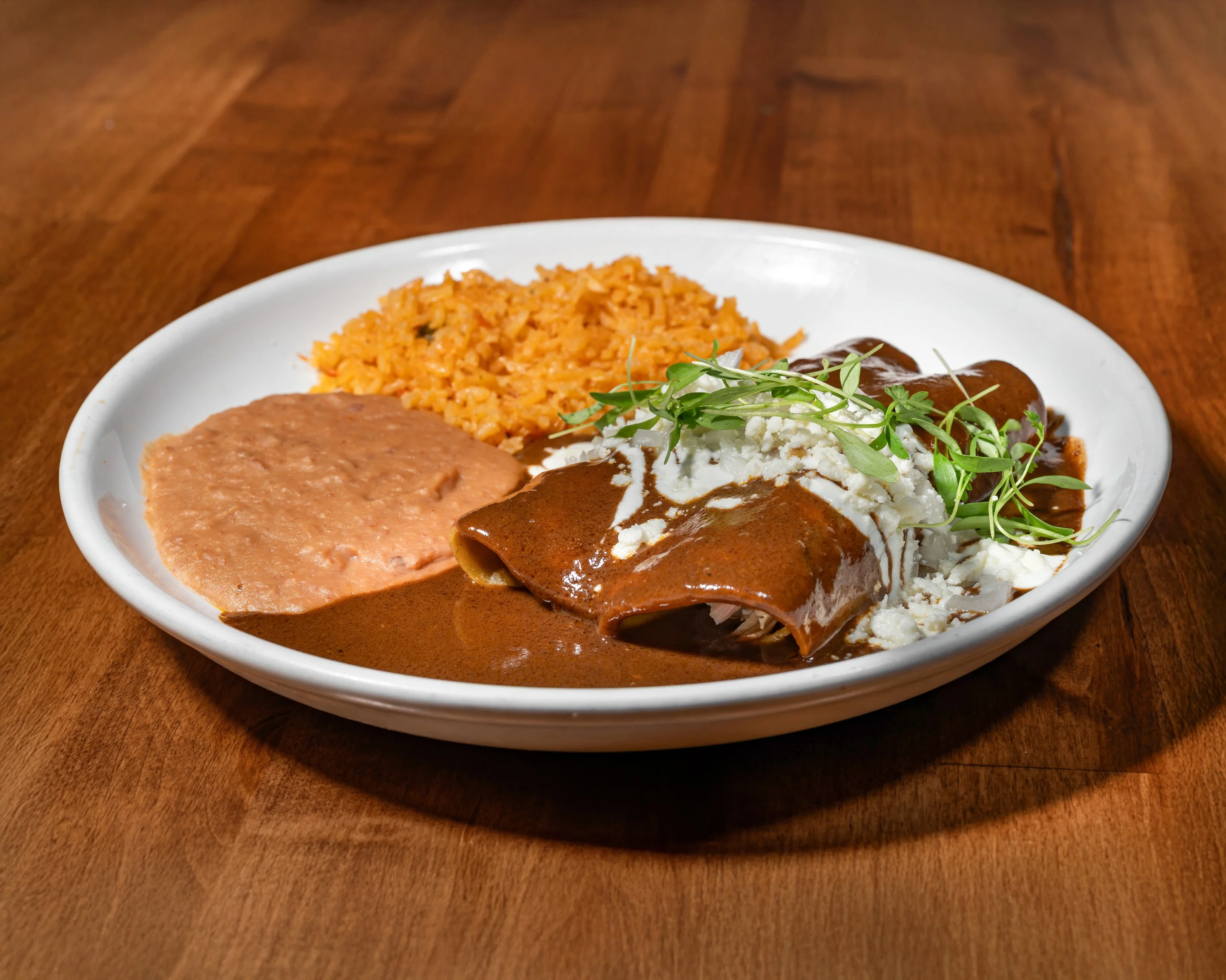 A plate featuring enchiladas smothered in sauce, accompanied by a serving of refried beans and seasoned rice, garnished with green onions.