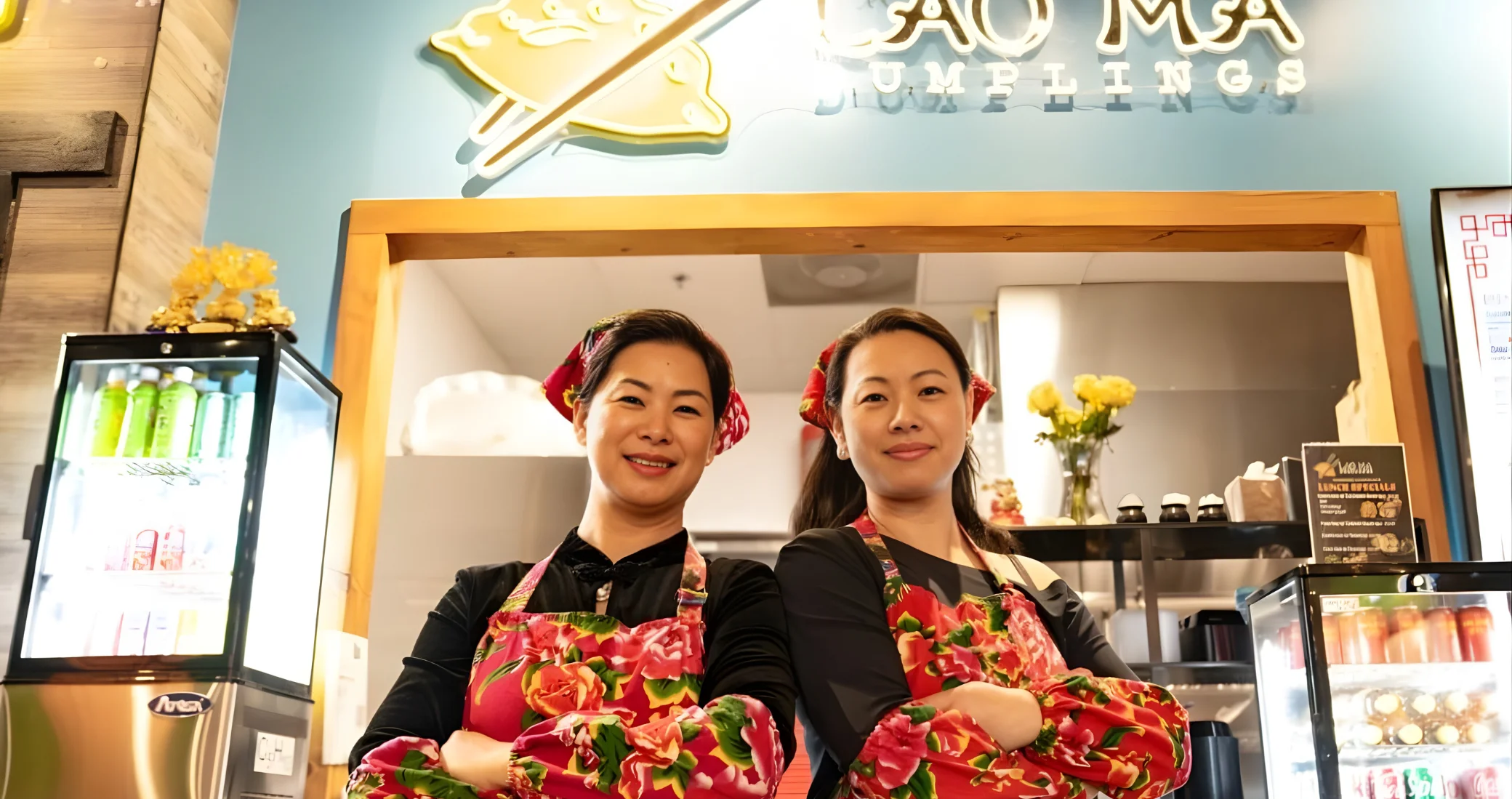 Two women in floral aprons smiling at a dumpling restaurant.