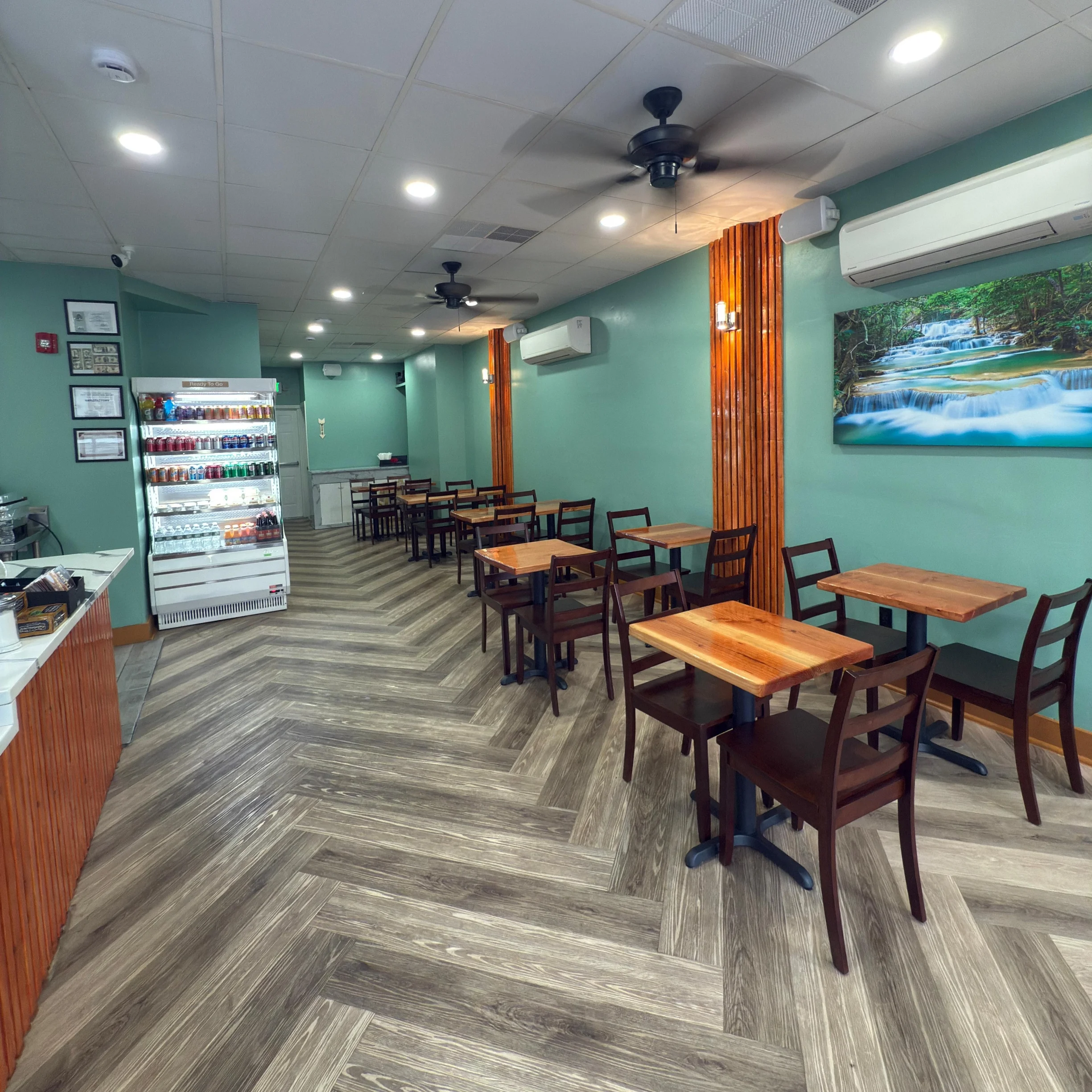 Small café interior with wooden tables, light green walls, decorative wooden slats, and a herringbone-pattern floor. A counter with drinks and snacks is on the left side, and a framed picture of a waterfall hangs on the right wall.