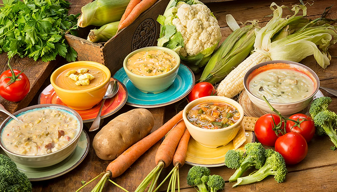 An inviting assortment of soups in colorful bowls, surrounded by fresh vegetables like carrots, tomatoes, corn, broccoli, and cauliflower on a rustic wooden table. The soups vary in color and texture, including creamy and broth-based varieties, evoking a farm-to-table dining experience.