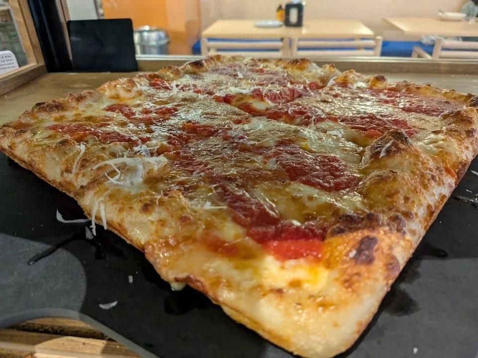 A square pizza with a crispy crust, topped with marinara sauce and melted cheese. The pizza is served on a black pizza tray.