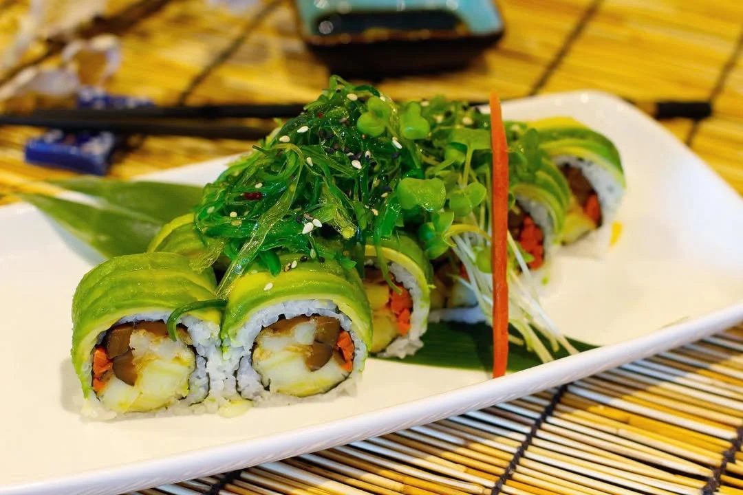 A sushi roll topped with avocado and garnished with green seaweed salad, presented on a white plate with a bamboo mat in the background.