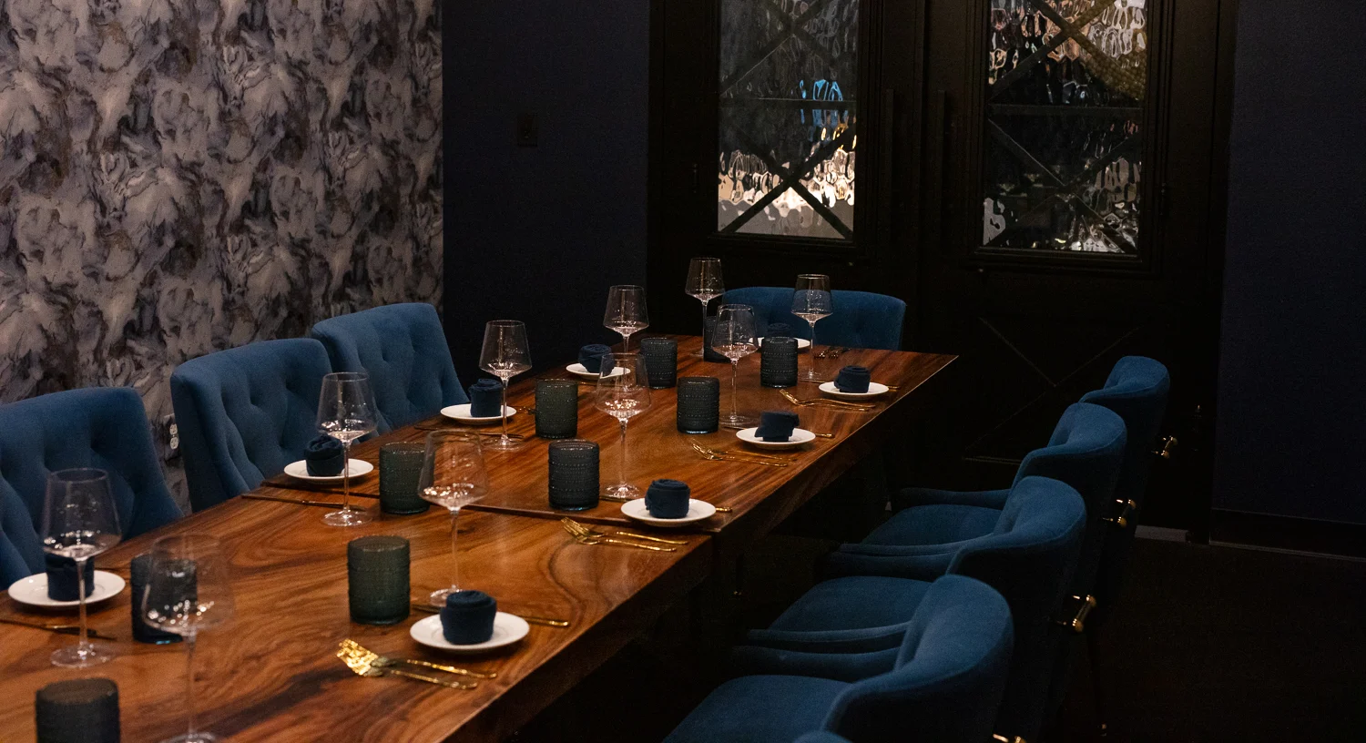 Elegant private dining room with a live-edge wooden table set for ten. Deep blue velvet chairs surround the table, which is arranged with gold flatware, navy napkins, modern water glasses, and crystal wine glasses. The space features a moody palette with textured gray marble wallpaper and dark double doors with frosted glass panels.