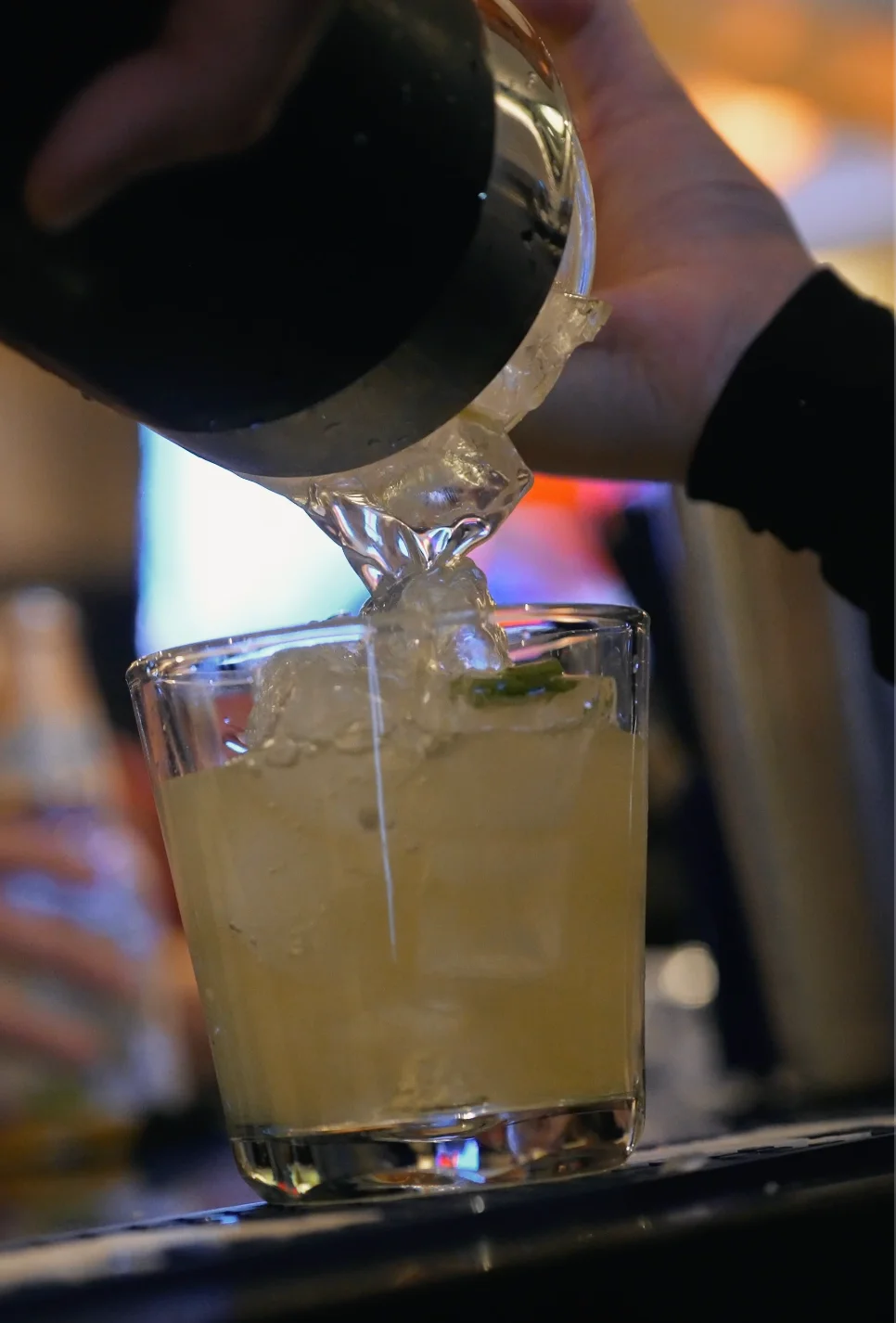 Bartender pouring a cocktail over ice in a glass.