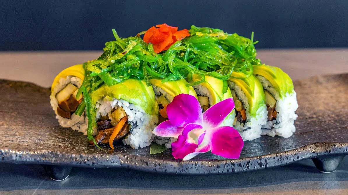 A sushi roll topped with avocado, garnished with seaweed and a decorative orchid. It showcases a blend of colorful ingredients, including carrots and sliced vegetables.