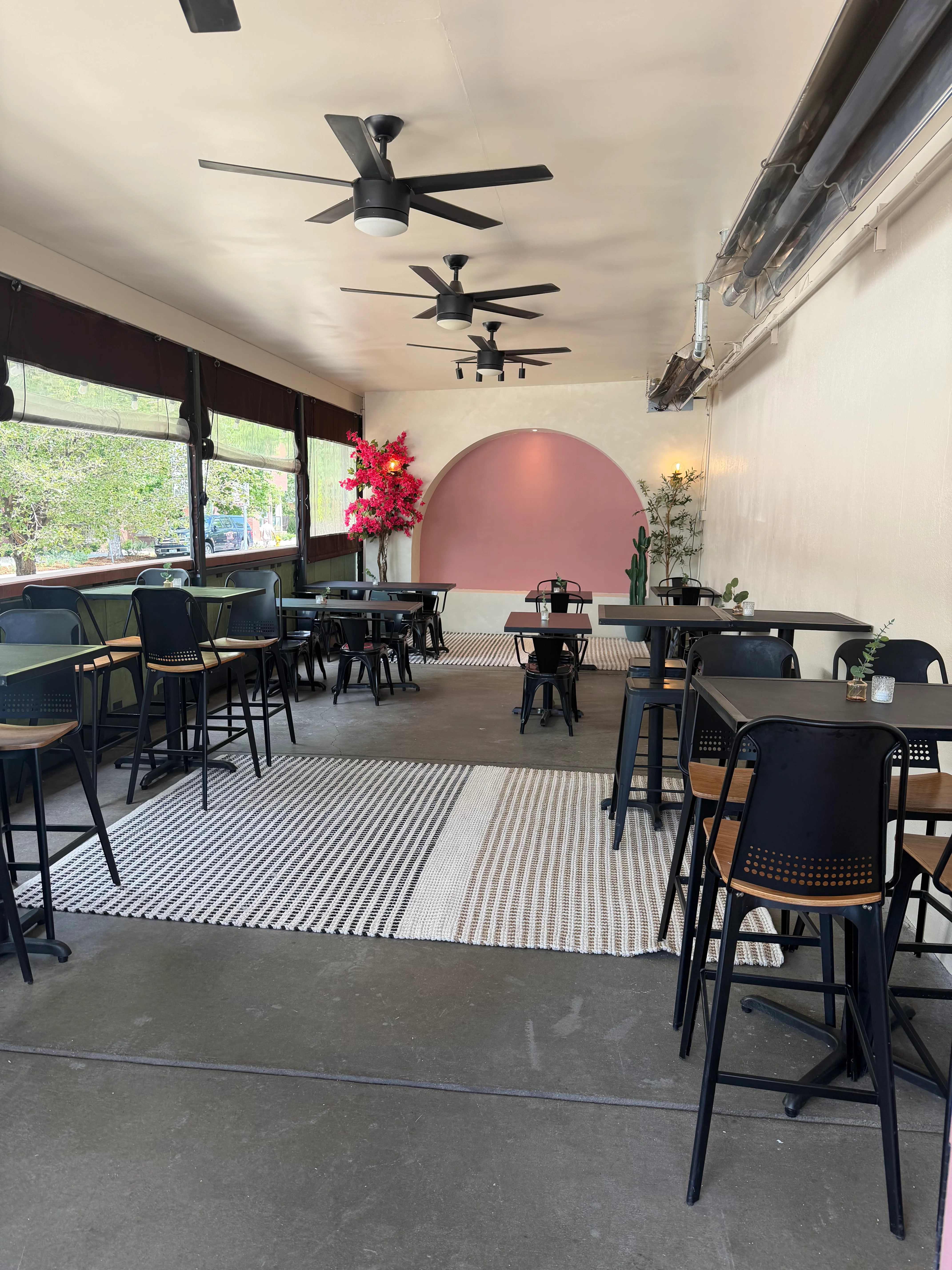 An indoor dining space featuring black tables and chairs, with ceiling fans and a decorative pink wall. A potted plant adds a touch of greenery.