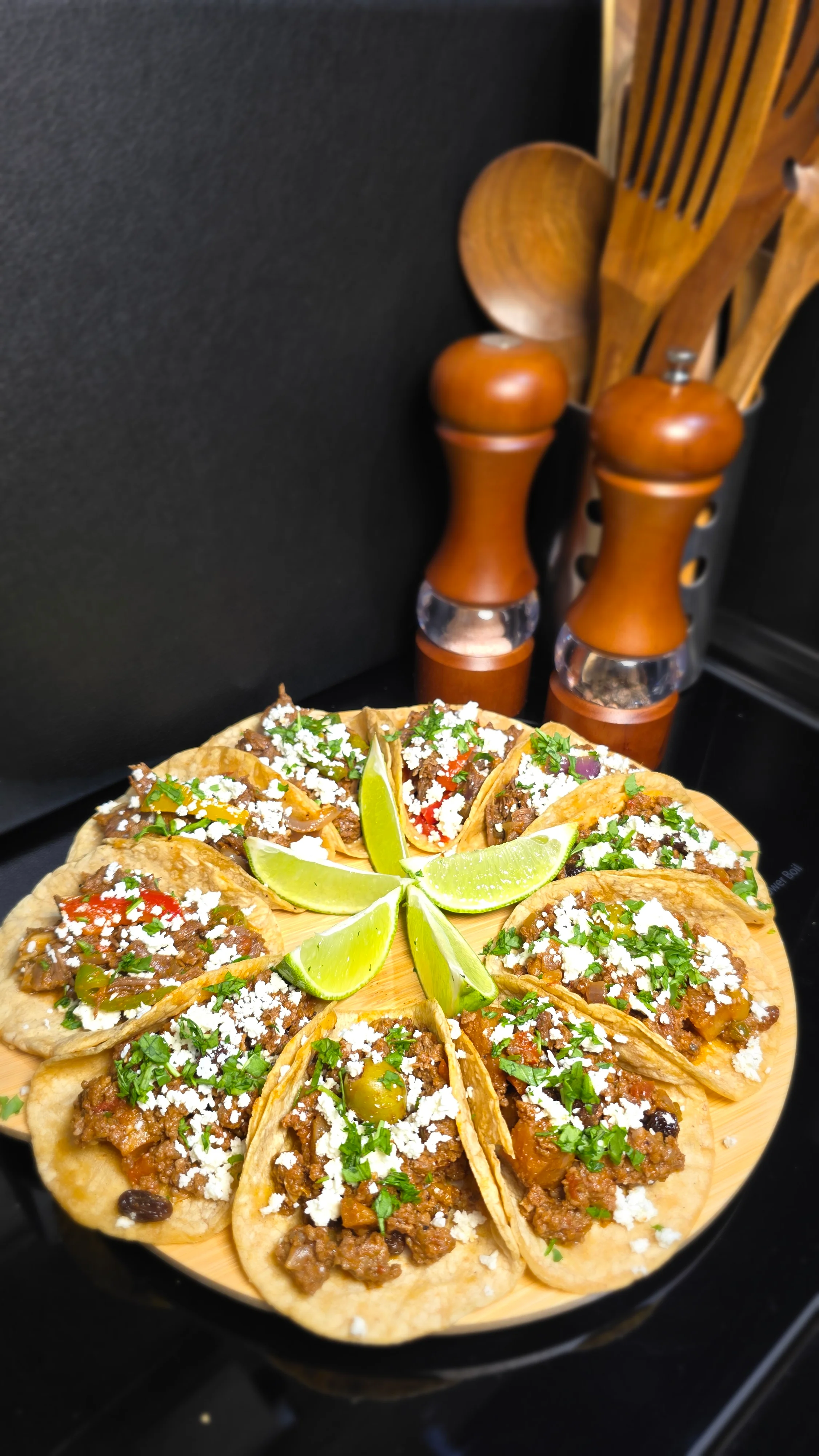 A beautifully arranged plate of tacos topped with various meats, onions, cilantro, and crumbled cheese, garnished with lime wedges, set against a dark background.