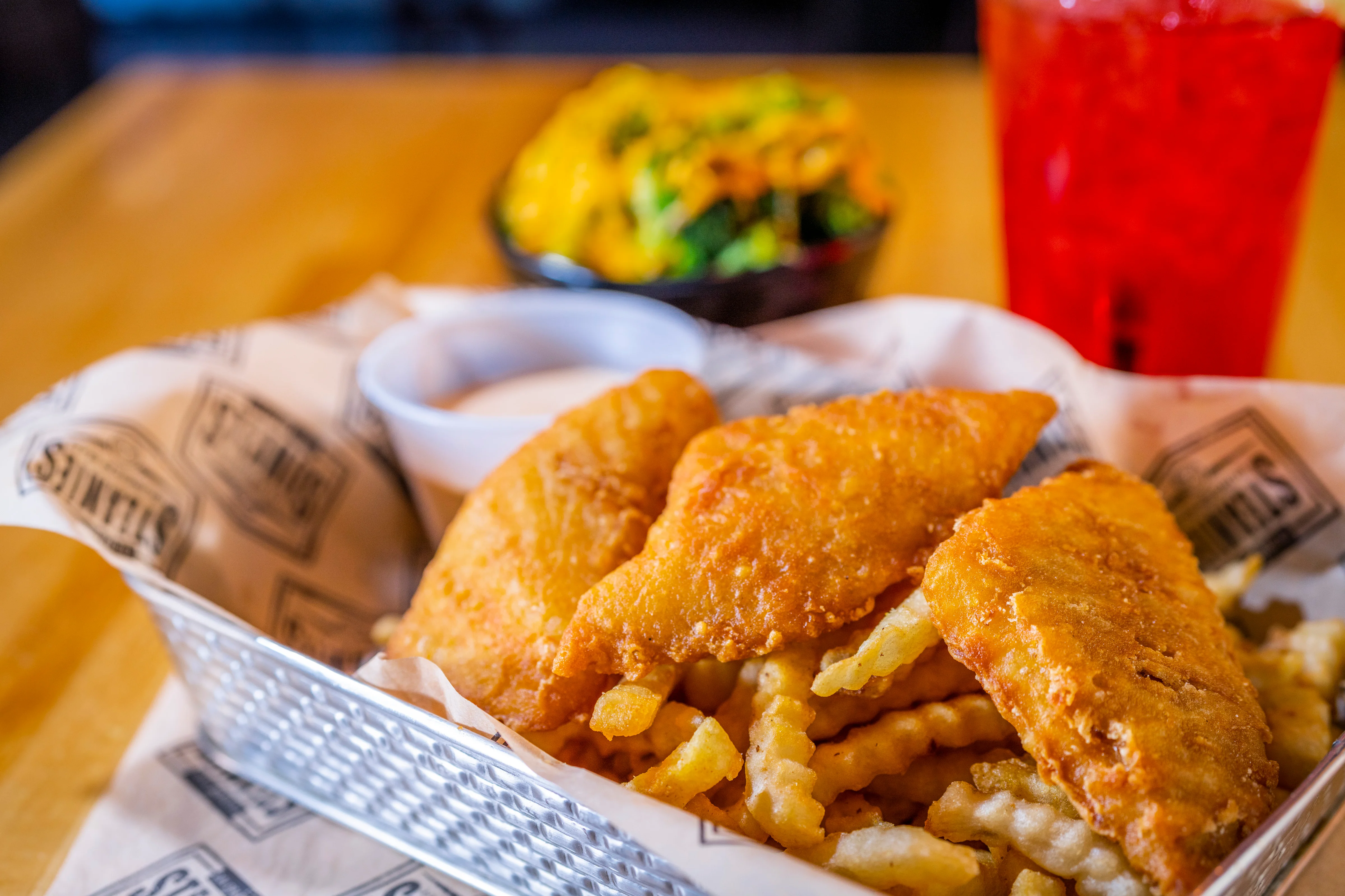 A basket of crispy fried fish served with crinkle cut fries and a side of dipping sauce, set on a rustic wooden table.
