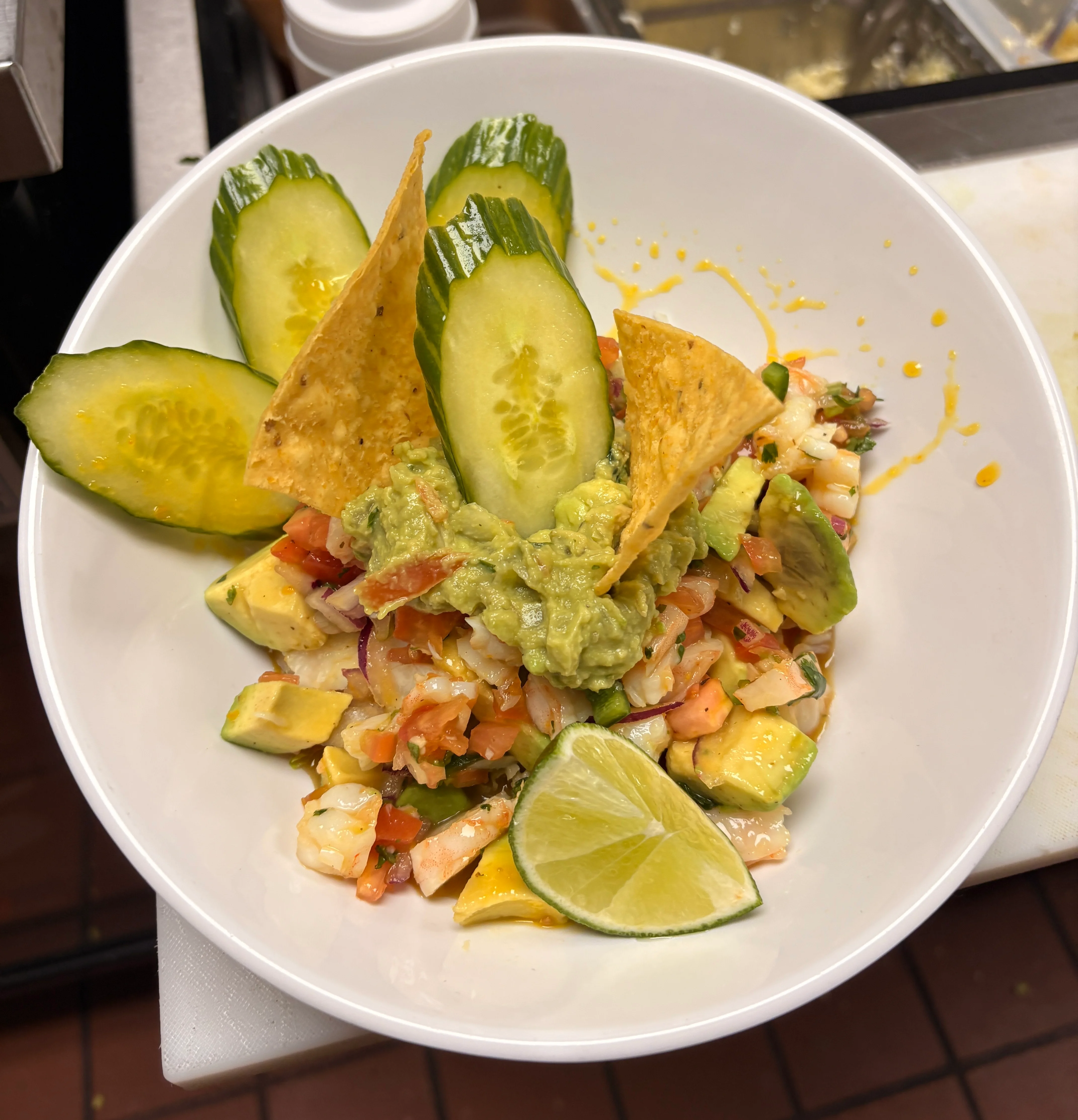 A vibrant dish featuring diced seafood, avocado, tomatoes, and guacamole, garnished with tortilla chips, cucumber slices, and a lime wedge.