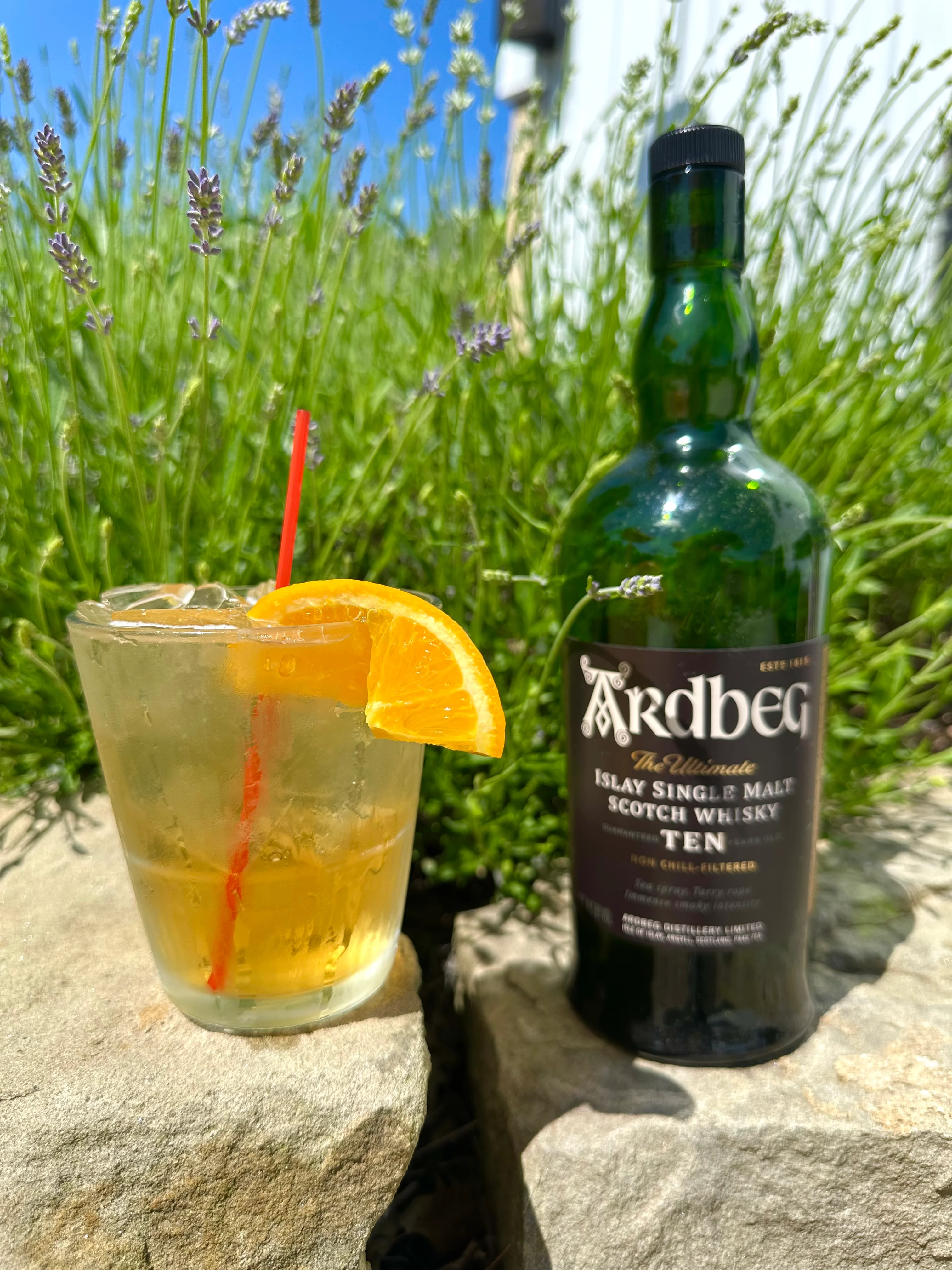 A glass of mixed drink garnished with an orange slice and a red straw, accompanied by a bottle of Islay single malt Scotch whisky, set against a backdrop of lavender plants.