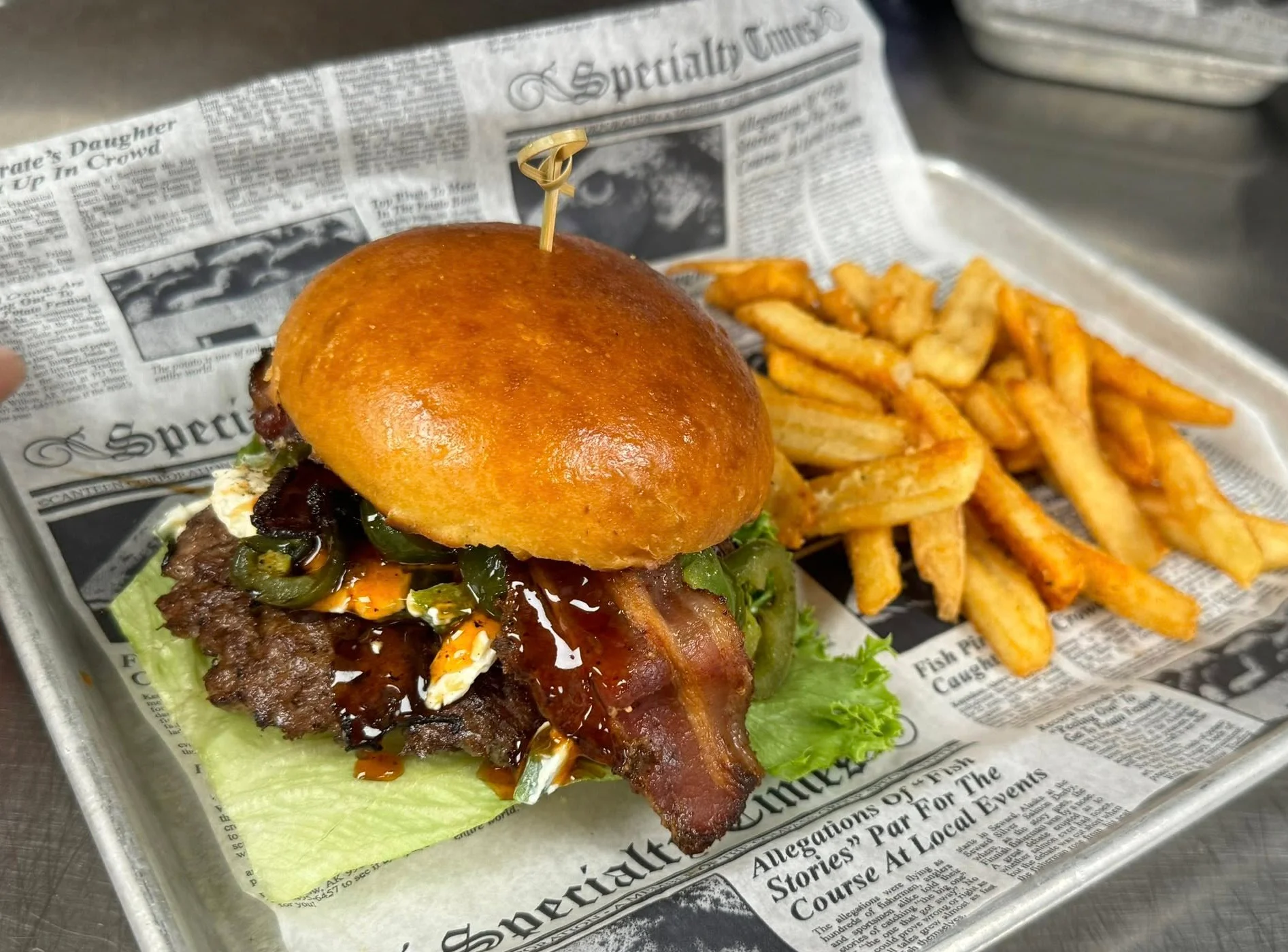 A bacon cheeseburger topped with jalapeños, lettuce, and barbecue sauce, served with fries on a tray lined with newspaper-style paper.
