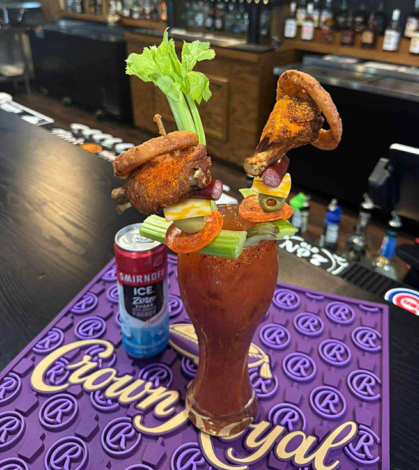 A tall Bloody Mary cocktail topped with celery, cheese cubes, olives, pepperoni, and fried chicken wings, served at the bar with liquor bottles in the background.