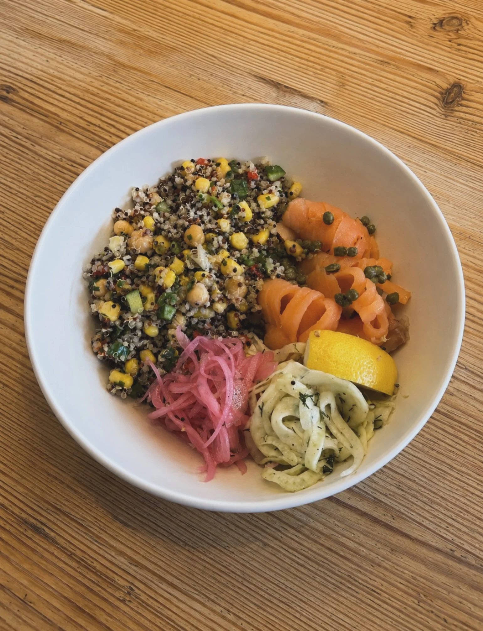 A bowl filled with quinoa, corn, smoked salmon, pickled onions, and lemon, arranged on a wooden table.