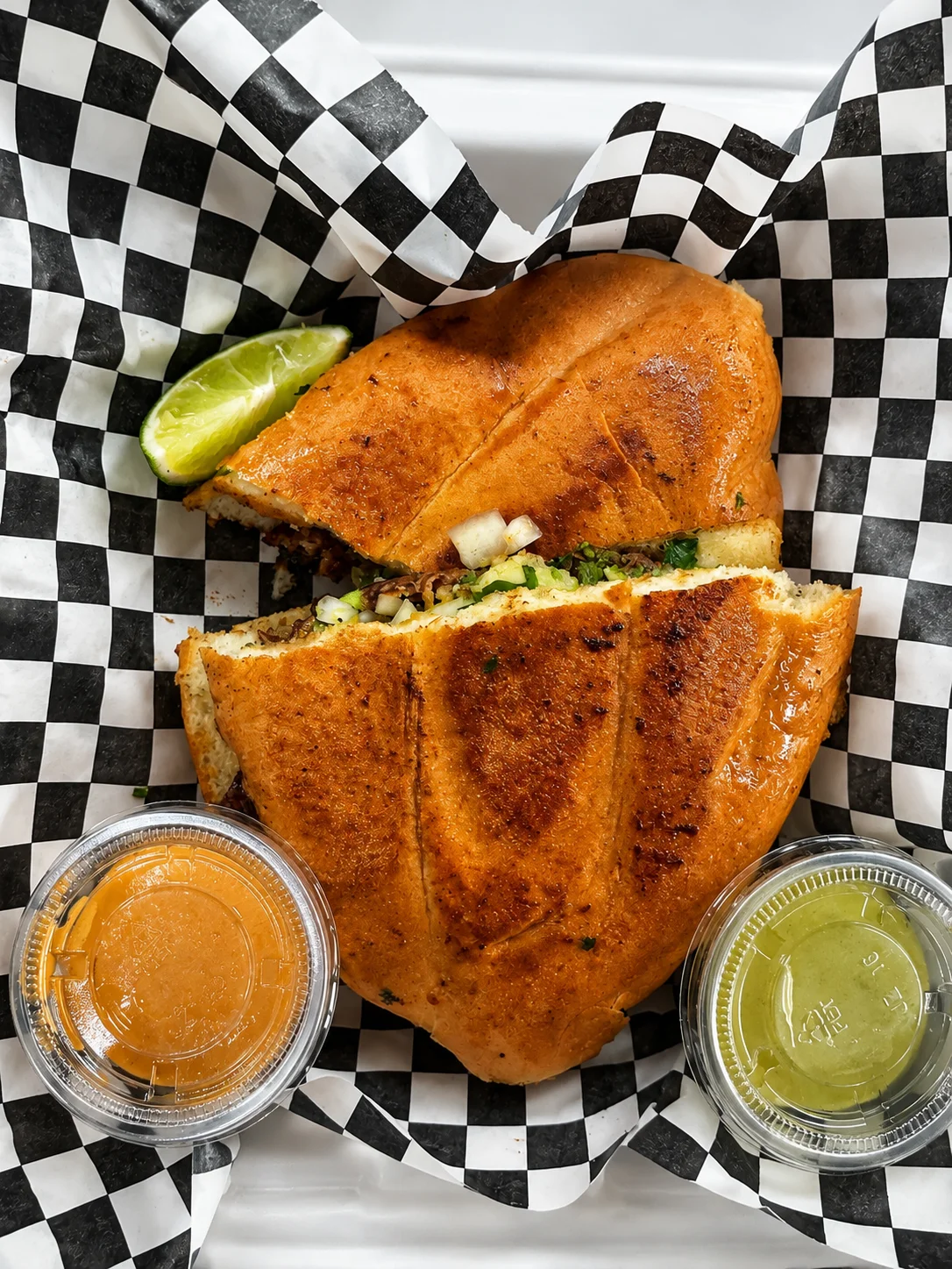 A halved sandwich served on a checkered paper, accompanied by lime and two small containers of sauce.