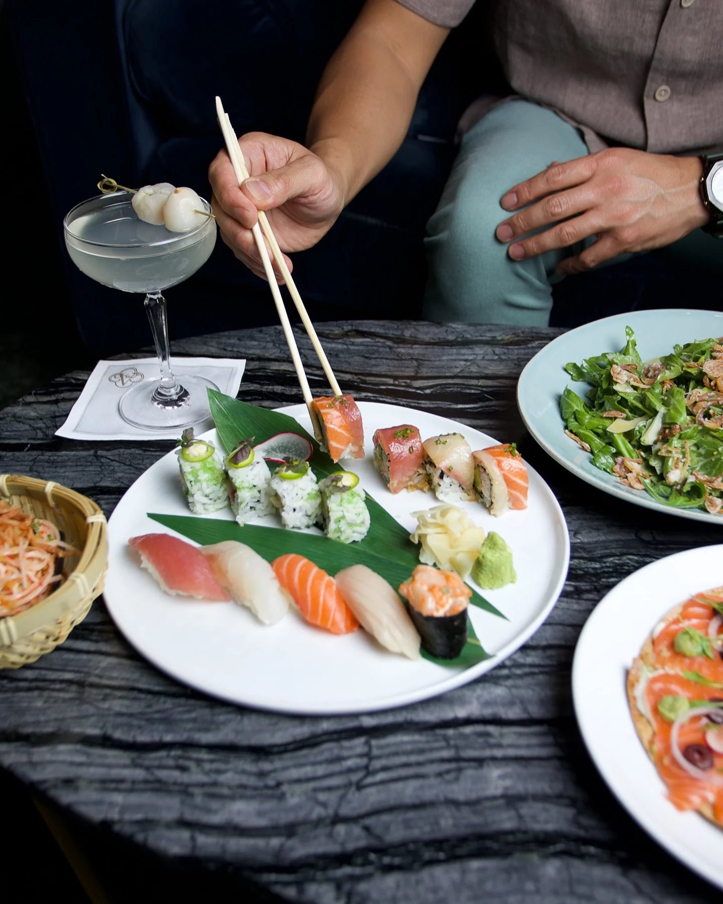 A person uses chopsticks to lift a sushi roll from a large white plate filled with assorted nigiri and specialty rolls, set on a dark, textured tabletop alongside a lychee-garnished cocktail, a green salad, and other dishes.