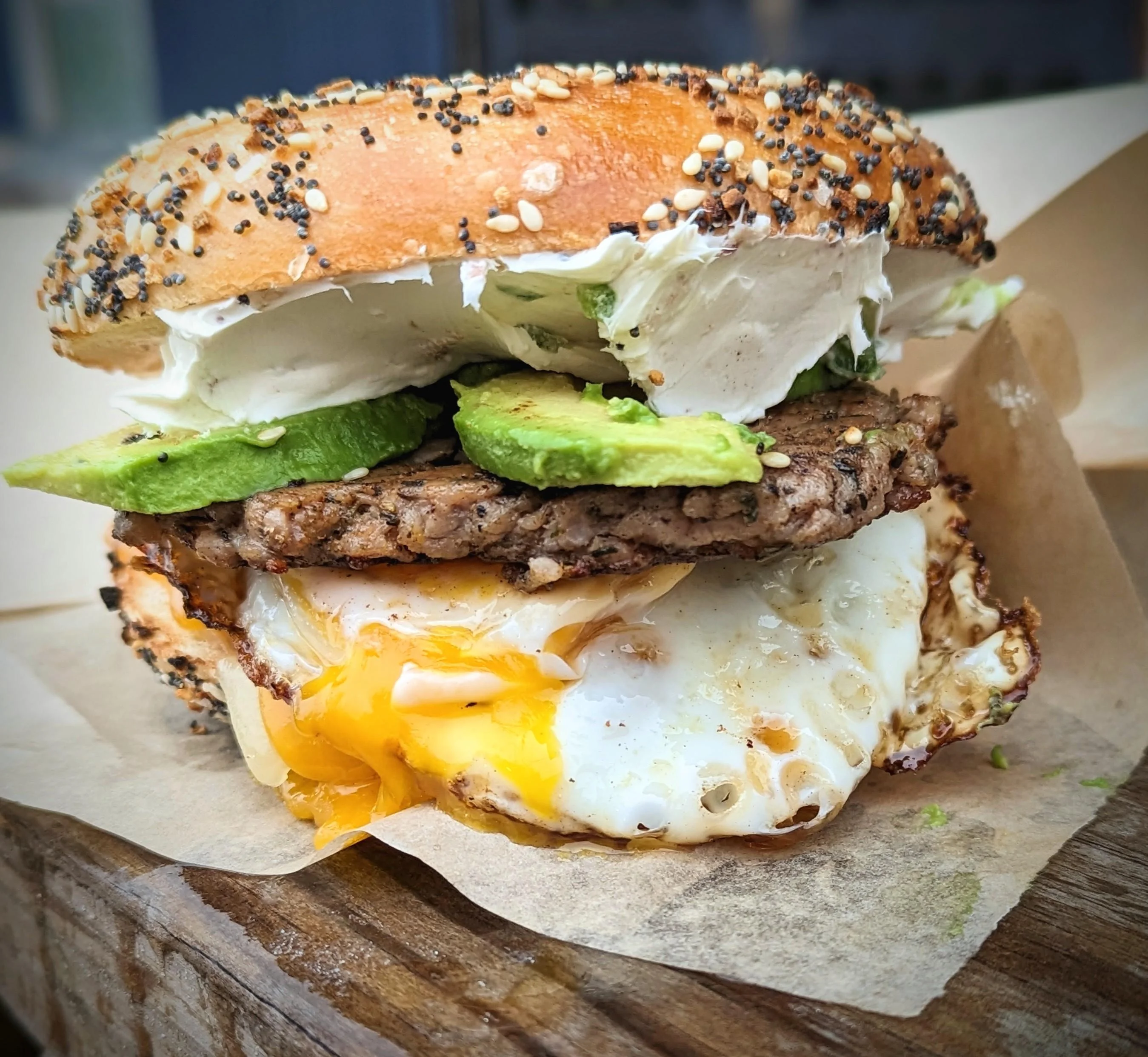 A hearty bagel sandwich stacked with a burger patty, fried egg, avocado, and cream cheese, presented on paper.