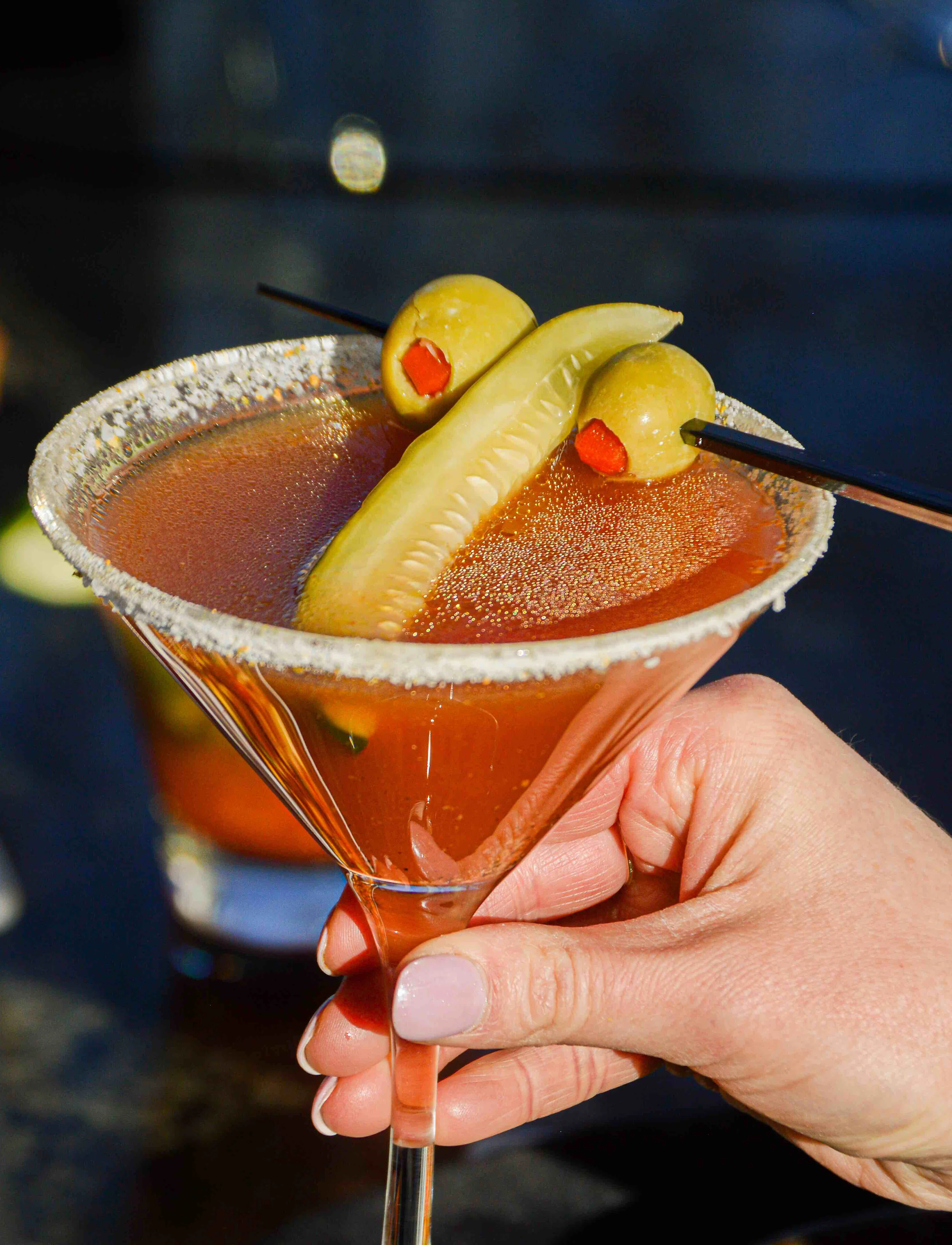 Naughty Pickle Martini is held in a room with a blue background, the cocktail has a pickle slice and 2 olives on the top with a salted rim