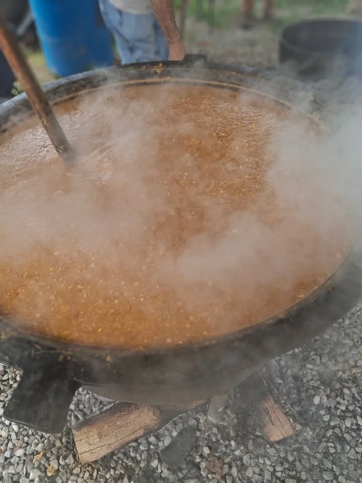 A large pot of steaming food being stirred outdoors, surrounded by a natural setting.