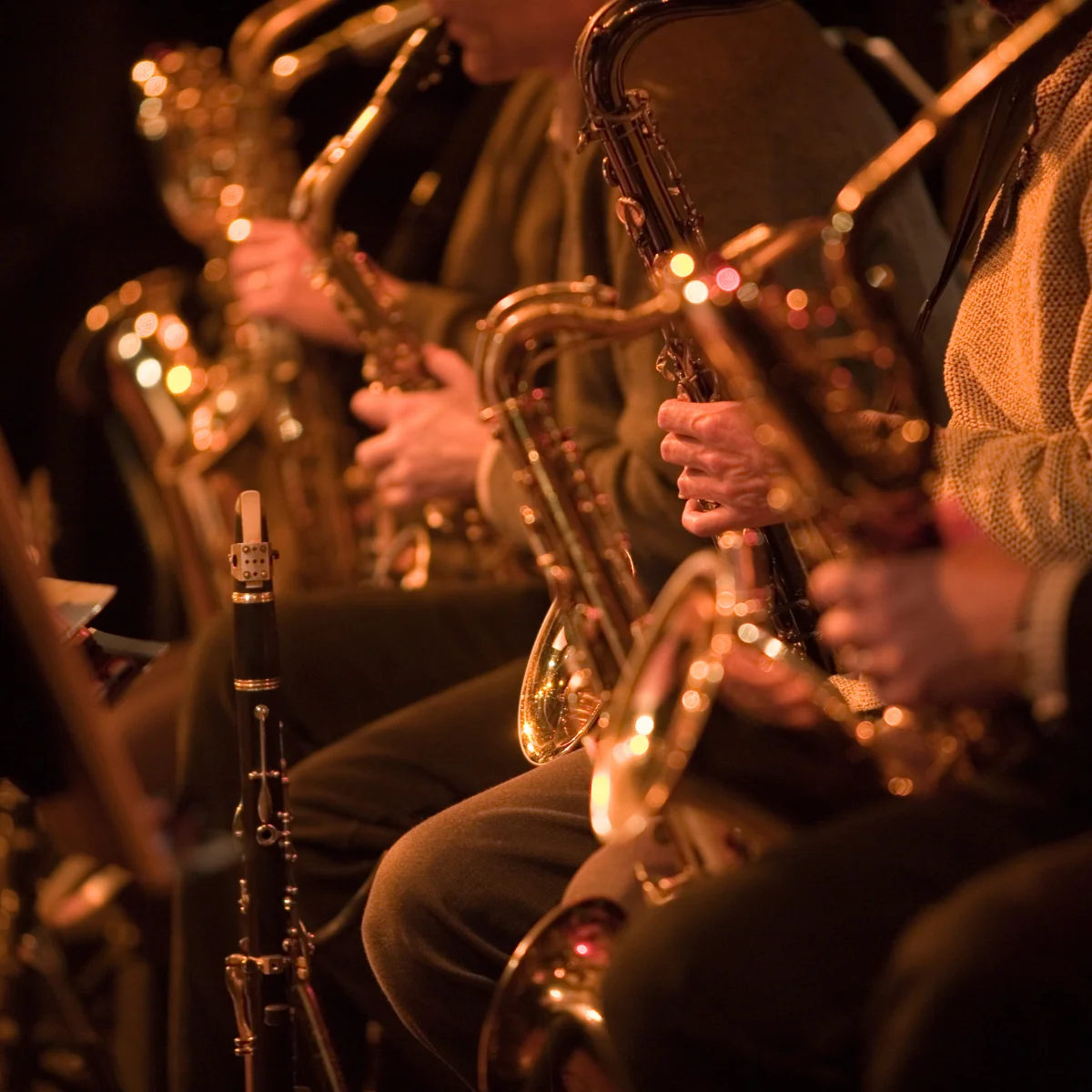Close-up of a jazz band’s saxophone section, with musicians in warm lighting holding shiny brass saxophones and a clarinet visible in the foreground.