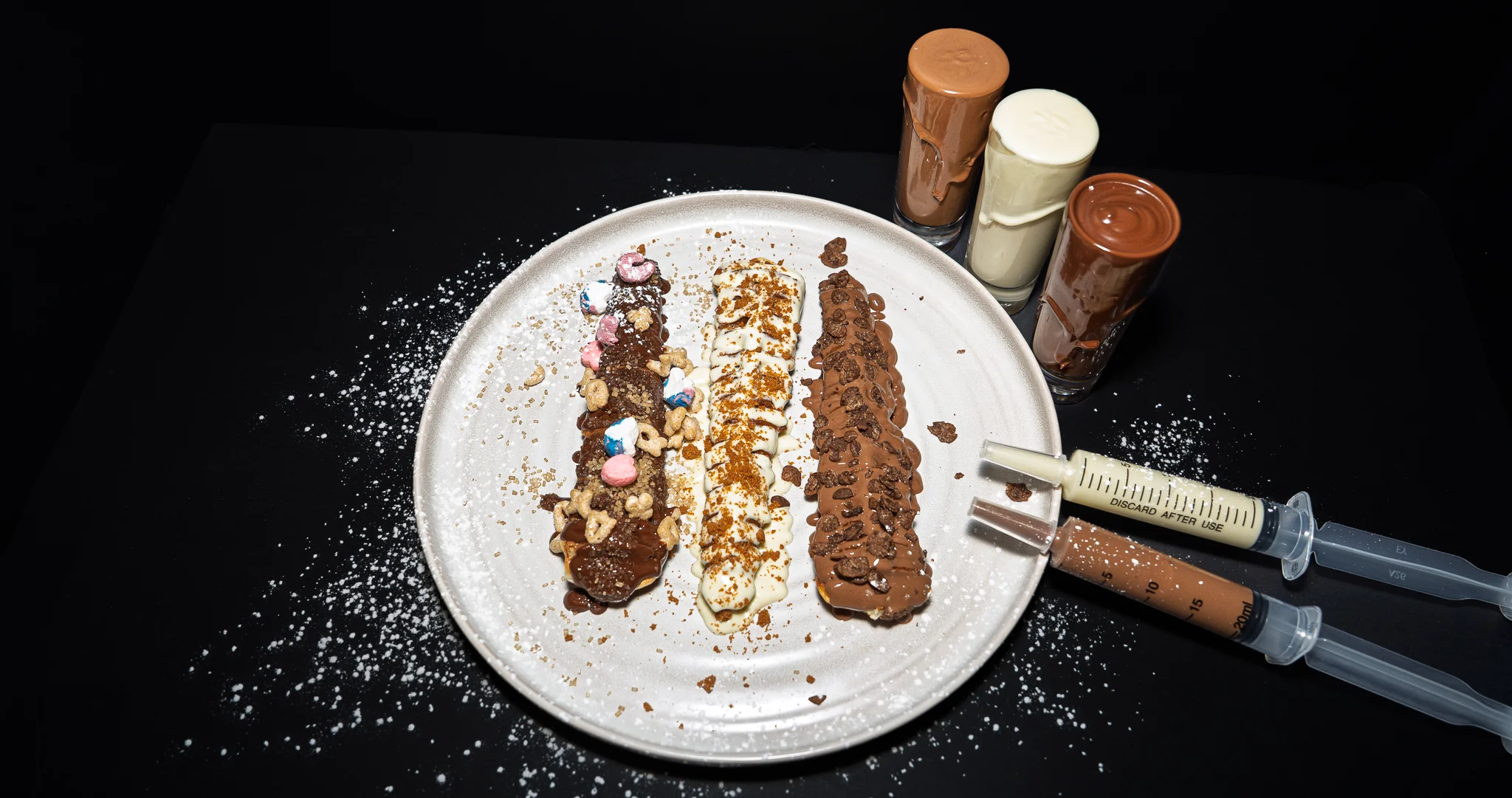 Three dessert rolls displayed on a black plate, featuring a combination of chocolate, caramel, and green toppings, all drizzled with white sauce and sprinkled with crunchy bits.