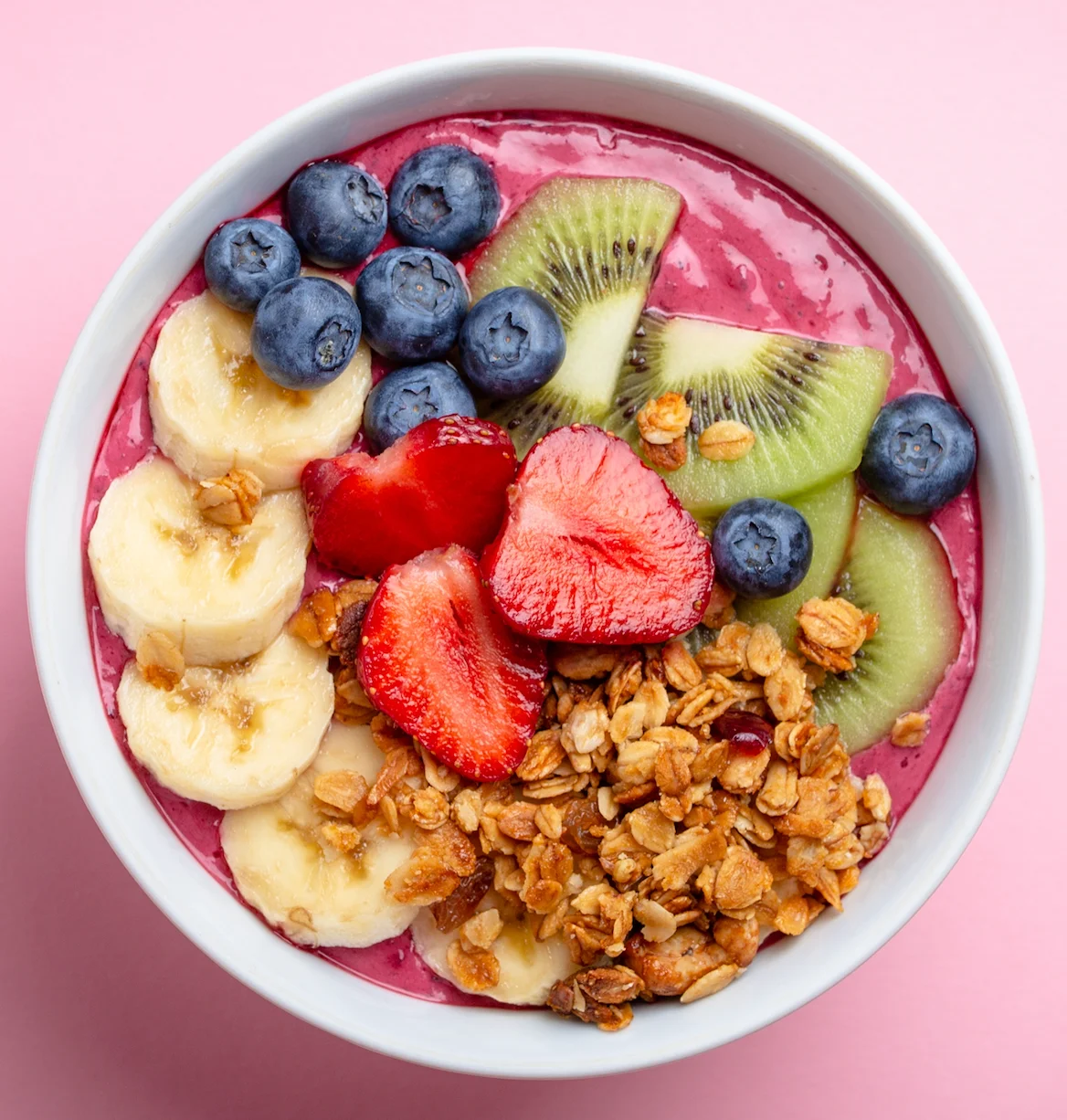 A vibrant smoothie bowl topped with banana, strawberries, blueberries, kiwi, and granola in a white bowl on a pink background.