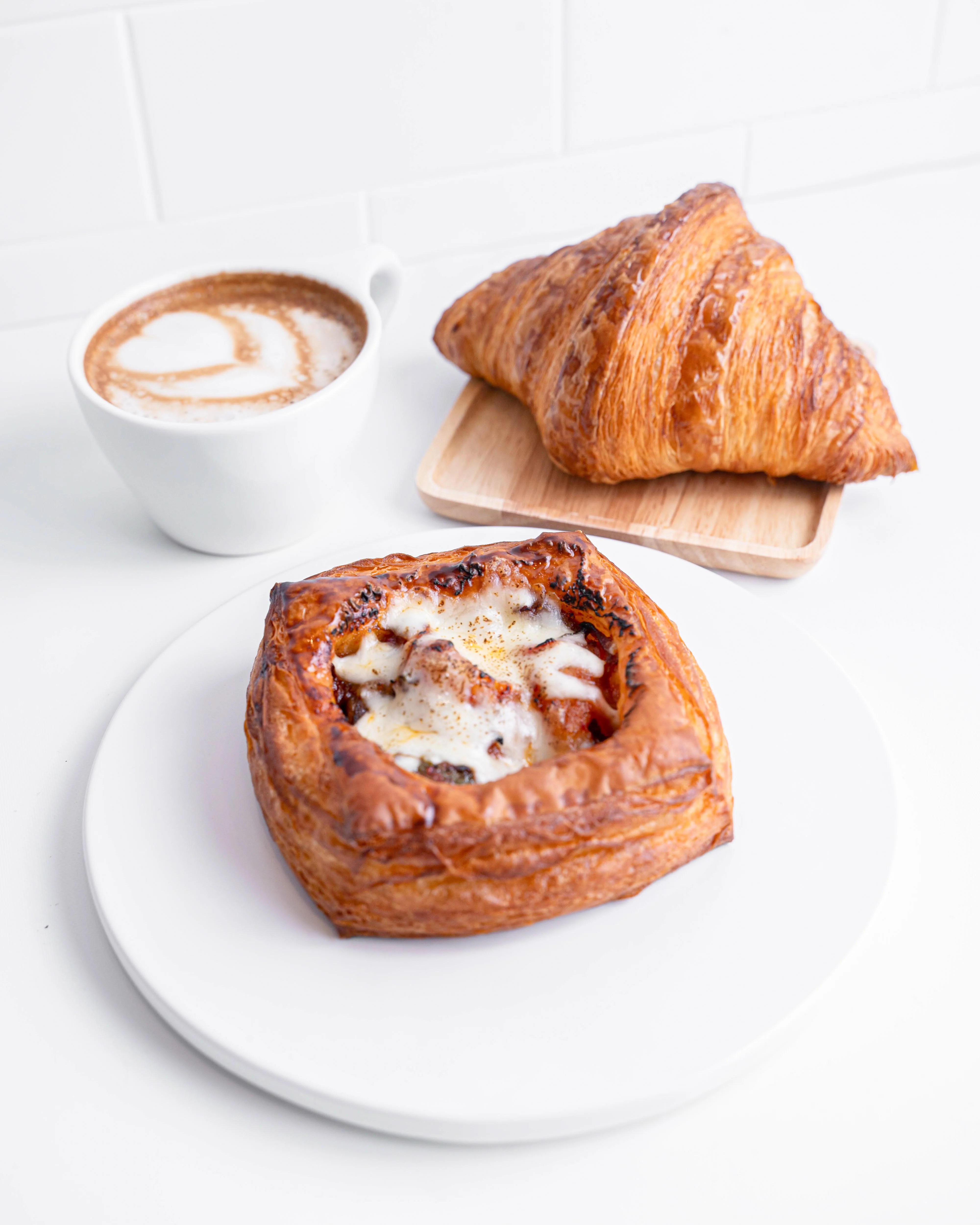A pastry filled with savory ingredients placed on a white plate next to a croissant and a cup of coffee with latte art.