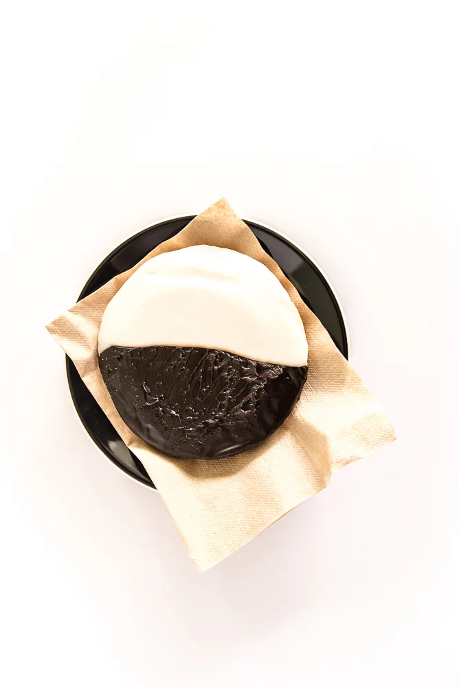 A black and white cookie, half chocolate, half vanilla glaze, on a beige napkin atop a black plate. The scene is minimalistic and inviting.