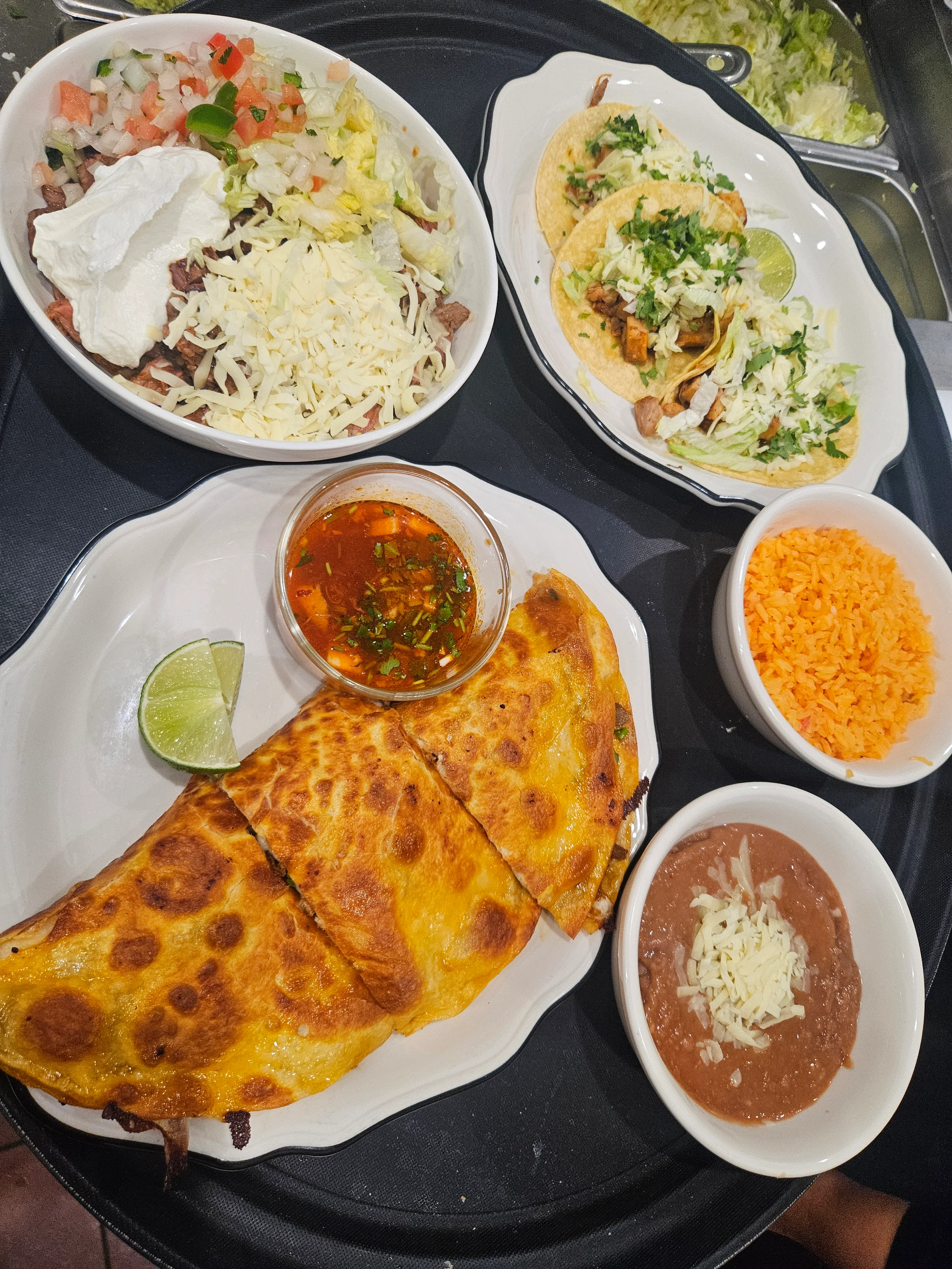 A colorful Mexican dish featuring a cheesy quesadilla, three tacos, rice, beans, and a salad with toppings. The meal is served with a side of sauce and lime.