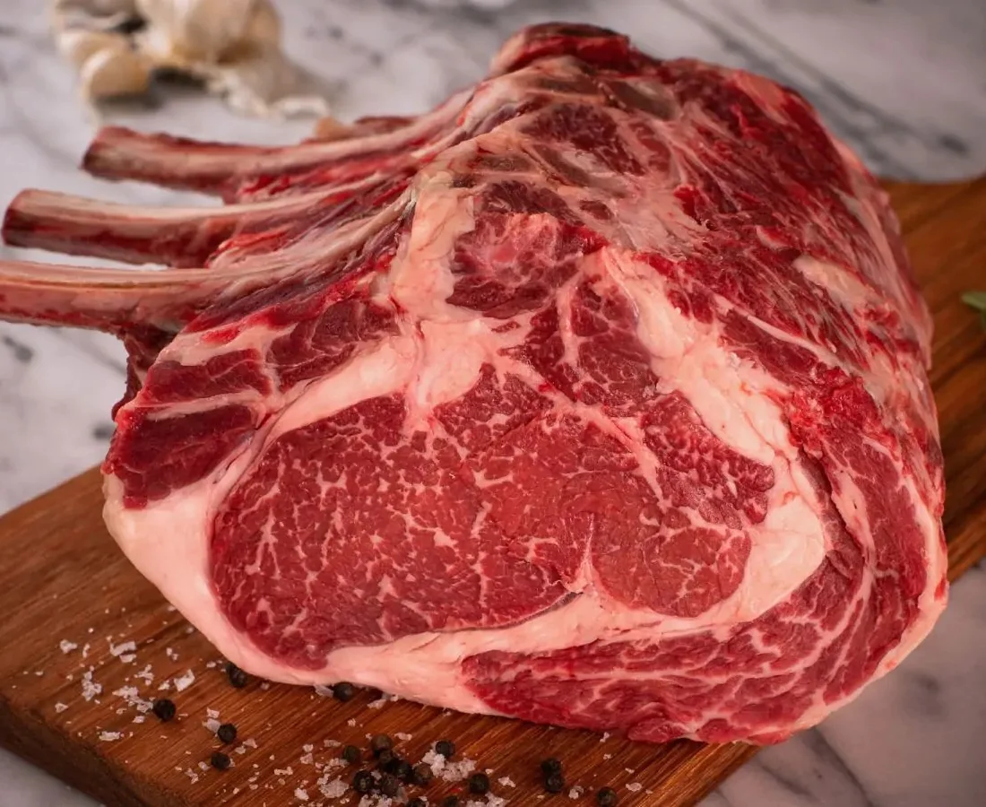 A close-up of a raw, bone-in standing rib roast resting on a wooden cutting board. The meat displays deep red muscle with extensive, intricate white marbling.