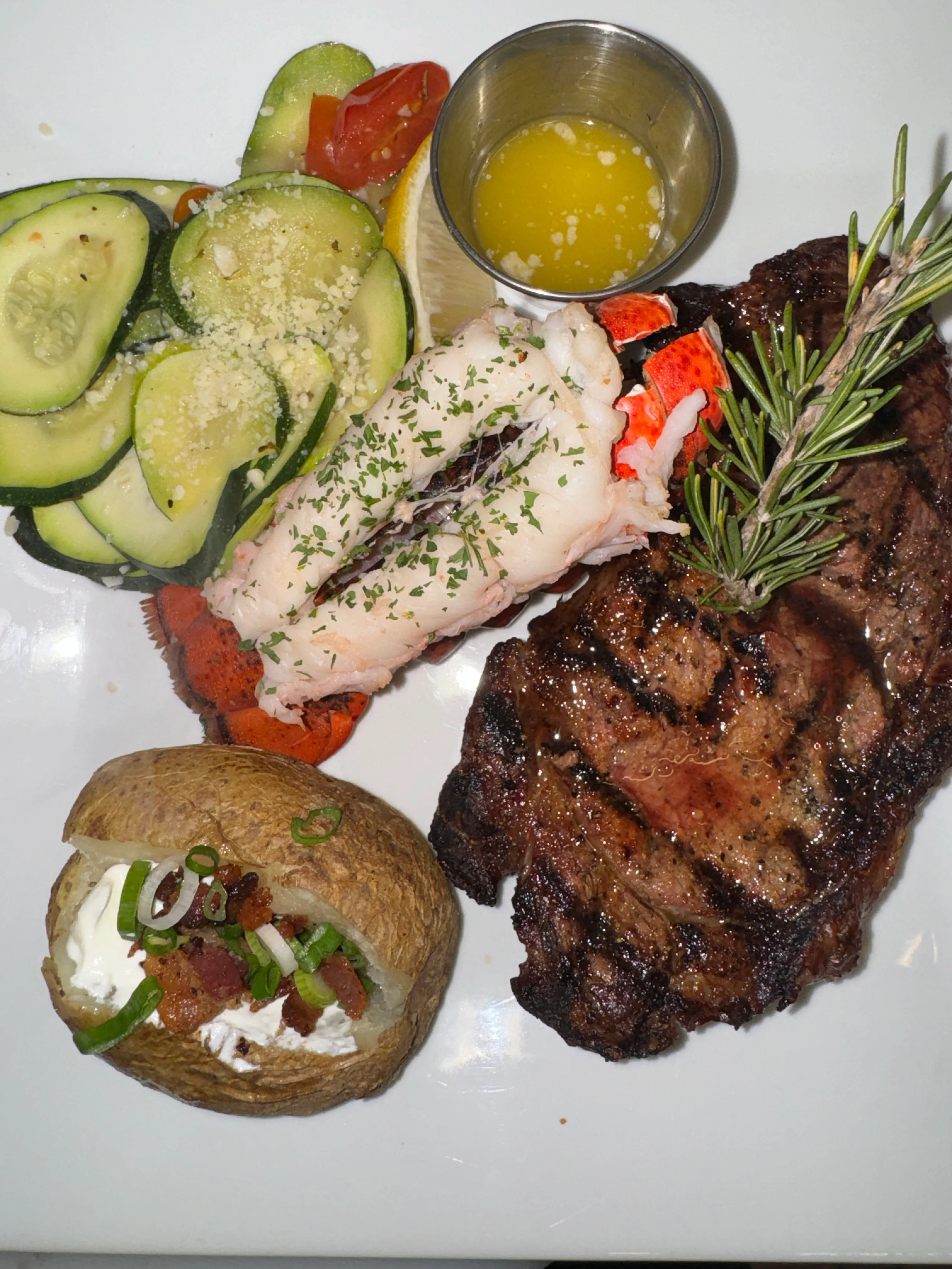 A hearty steak dinner complemented by grilled lobster, cucumber salad, and a baked potato with toppings.