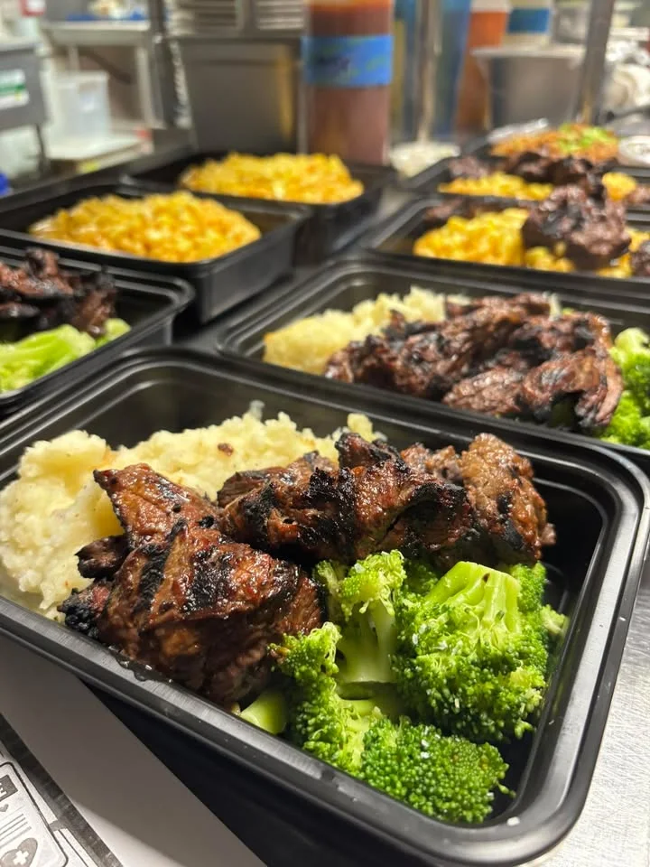 A meal prep setup featuring grilled meat alongside mashed potatoes and broccoli, presented in black containers.