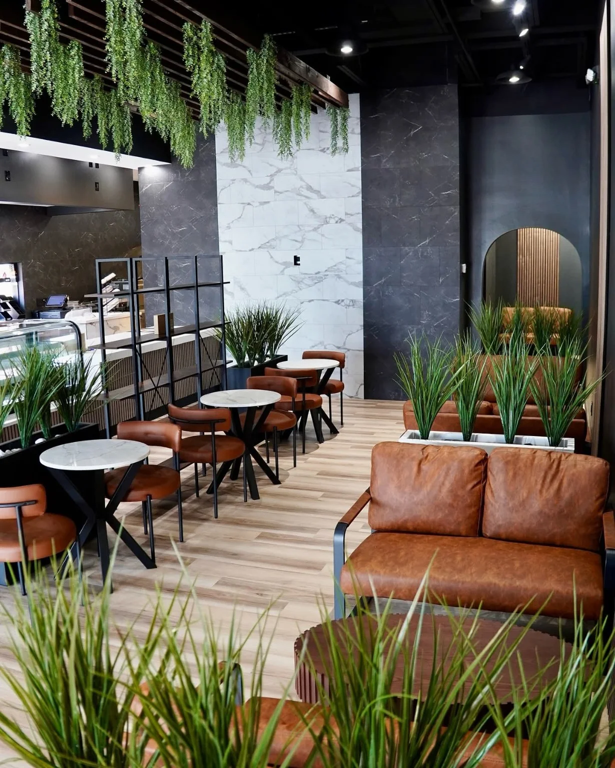 An inviting coffee shop layout with hanging plants, brown leather chairs, and small tables. The walls are finished in a sleek dark tone, adding to the contemporary atmosphere.