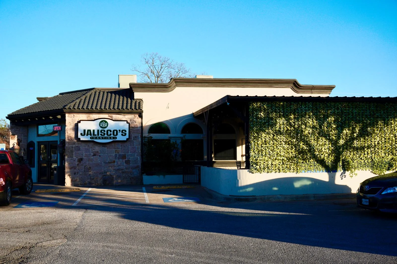 An exterior view of a restaurant named Jalisco's Cantina, featuring a stone facade, a green shrub wall, and a clear blue sky.