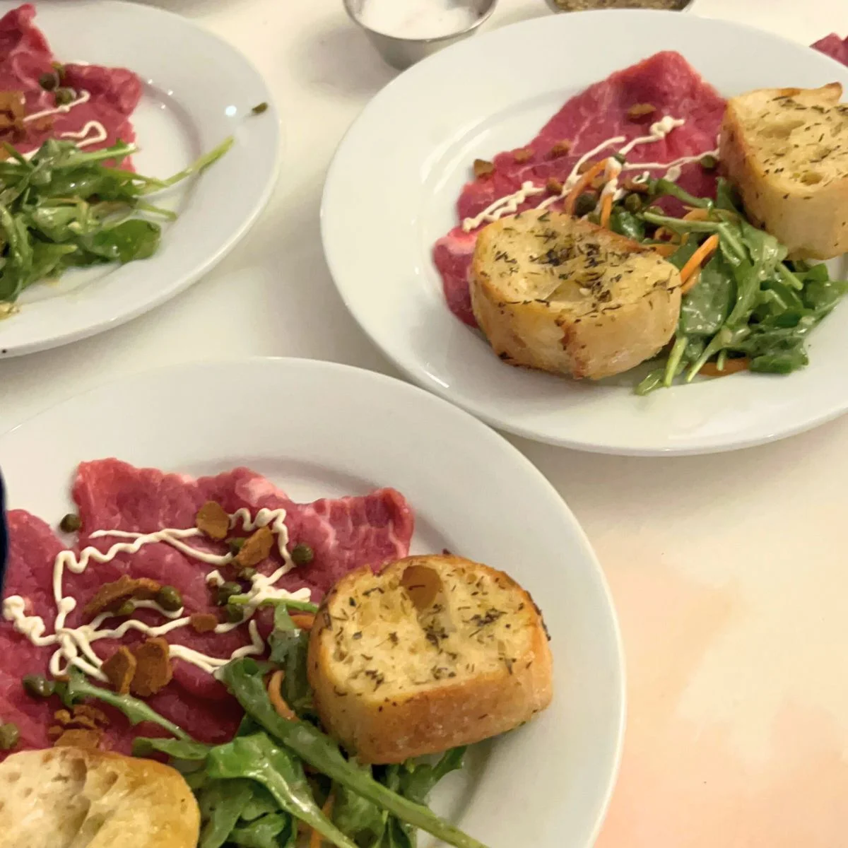Plates of carpaccio topped with sauce and capers, served with arugula salad and a slice of garlic bread.