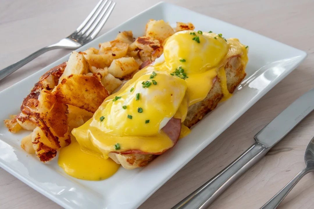 A plate of eggs Benedict topped with creamy hollandaise sauce, served on toasted bread with a side of golden, crispy hash browns.