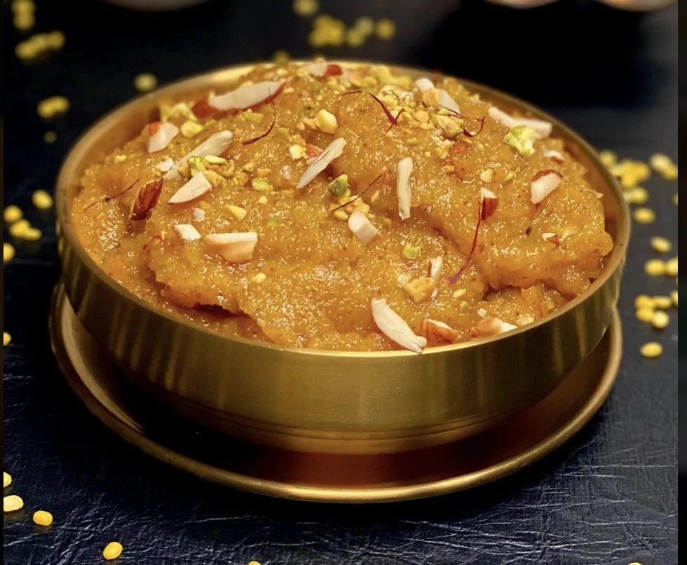 A golden colored Indian sweet dish garnished with nuts and saffron, served in a metallic bowl.