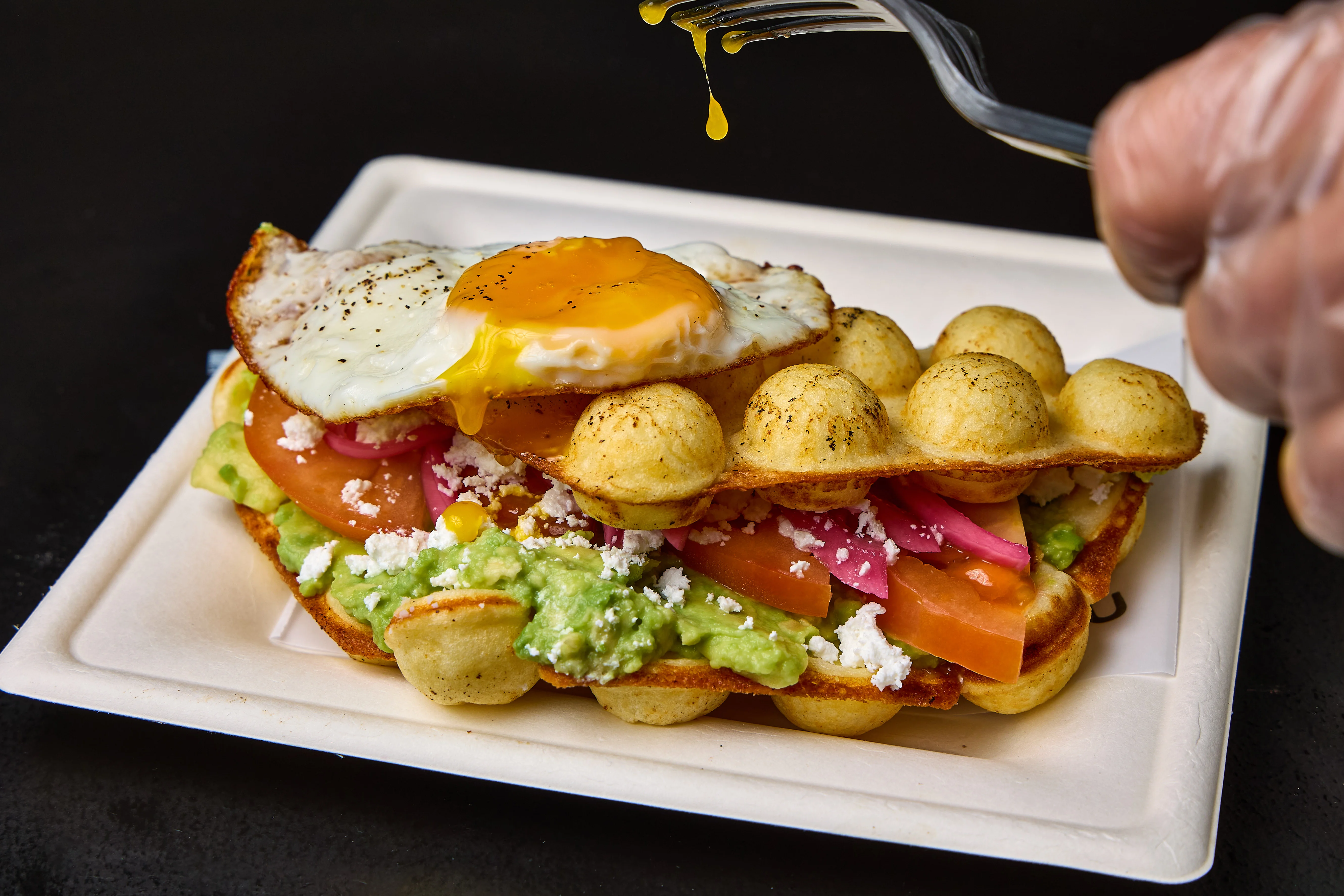 A delicious dish featuring a fried egg on top of a crispy base with layers of avocado, tomato, and cheese, highlighted by a drizzle of yolk.
