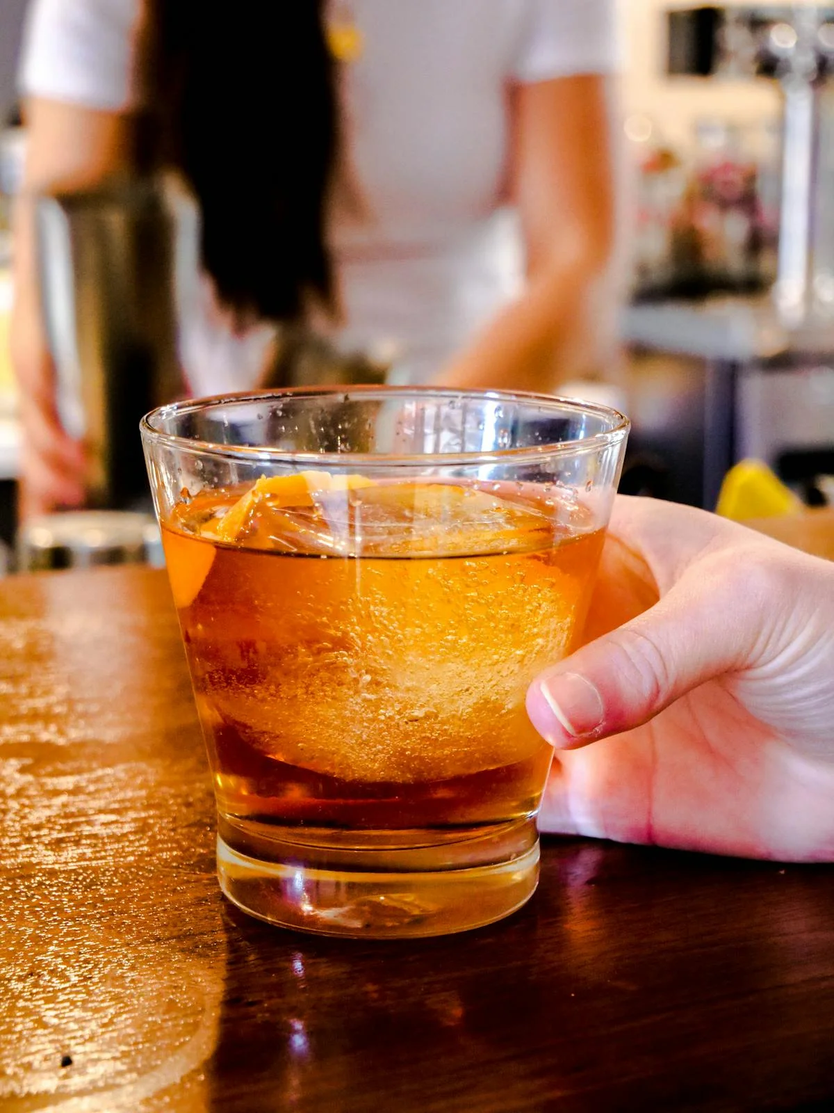 A hand holding a glass of amber-colored drink with ice and lemon slice.